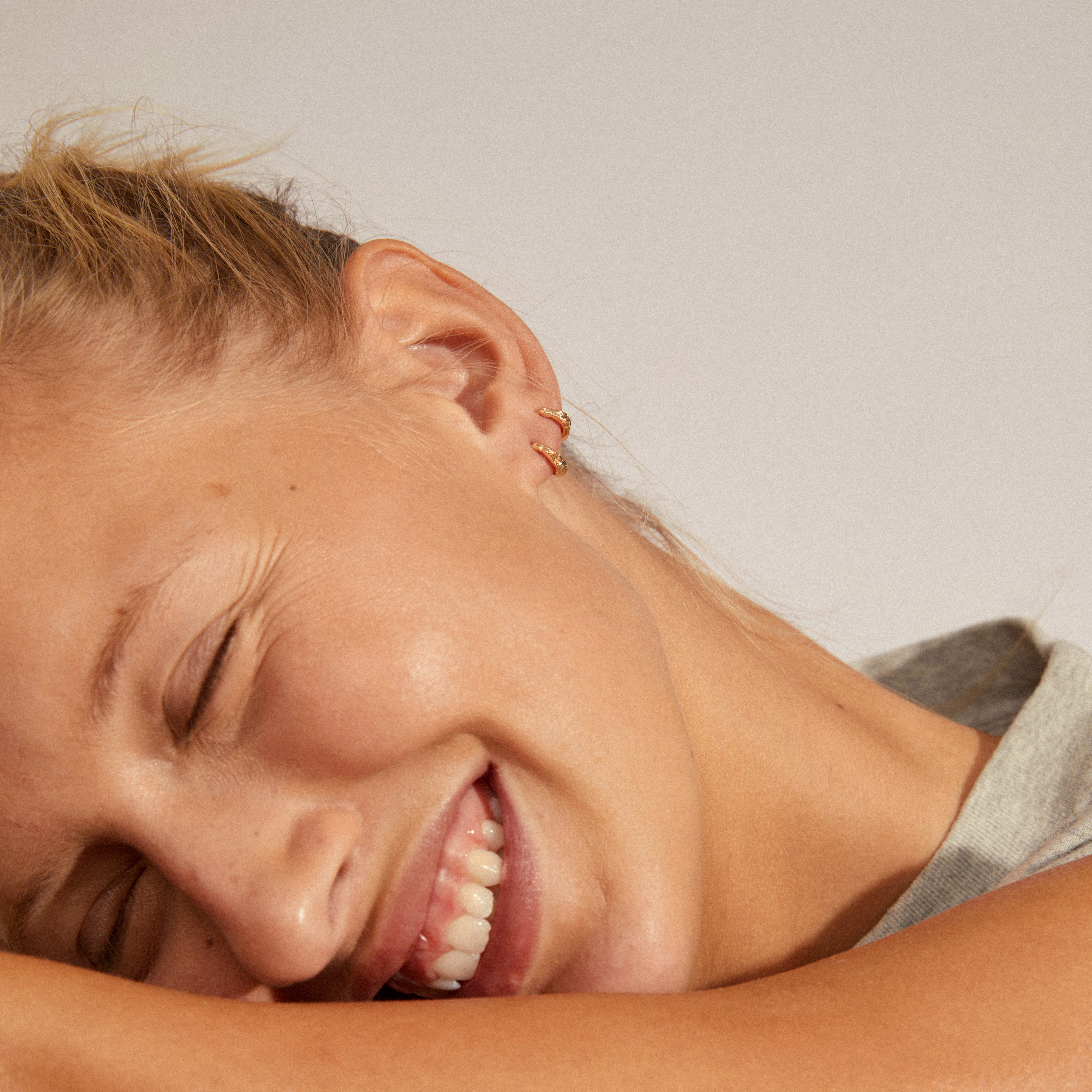 Woman wearing EDDY organic shaped mini huggie hoops gold-plated, eyes closed, smiling, showcasing the earrings' handcrafted and undulating design. Perfect for a curated ear look.