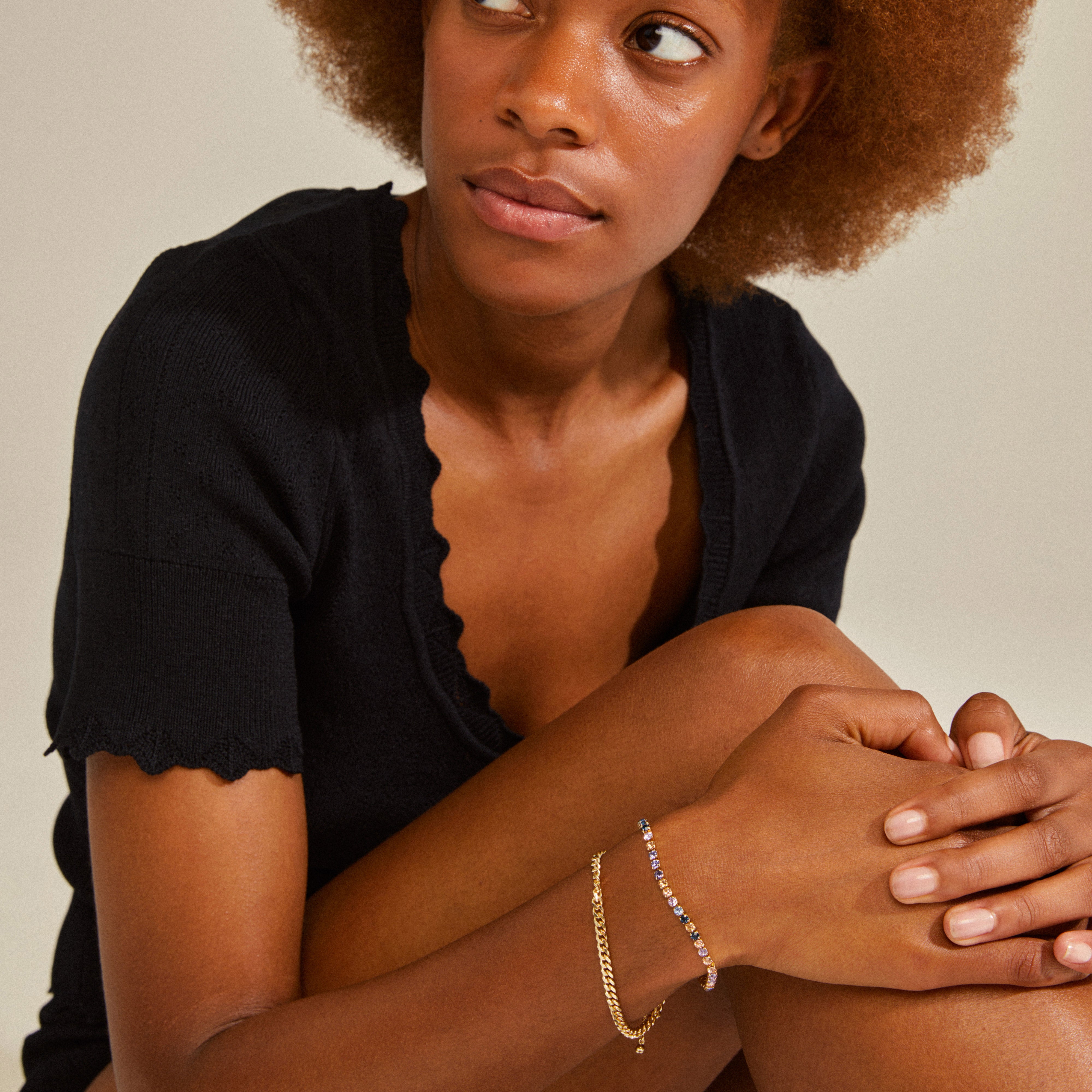 Woman wearing Pilgrim's REIGN bracelet, 2-in-1 set, gold-plated with curb chain and blue and champagne Preciosa crystals, sitting casually on the floor.