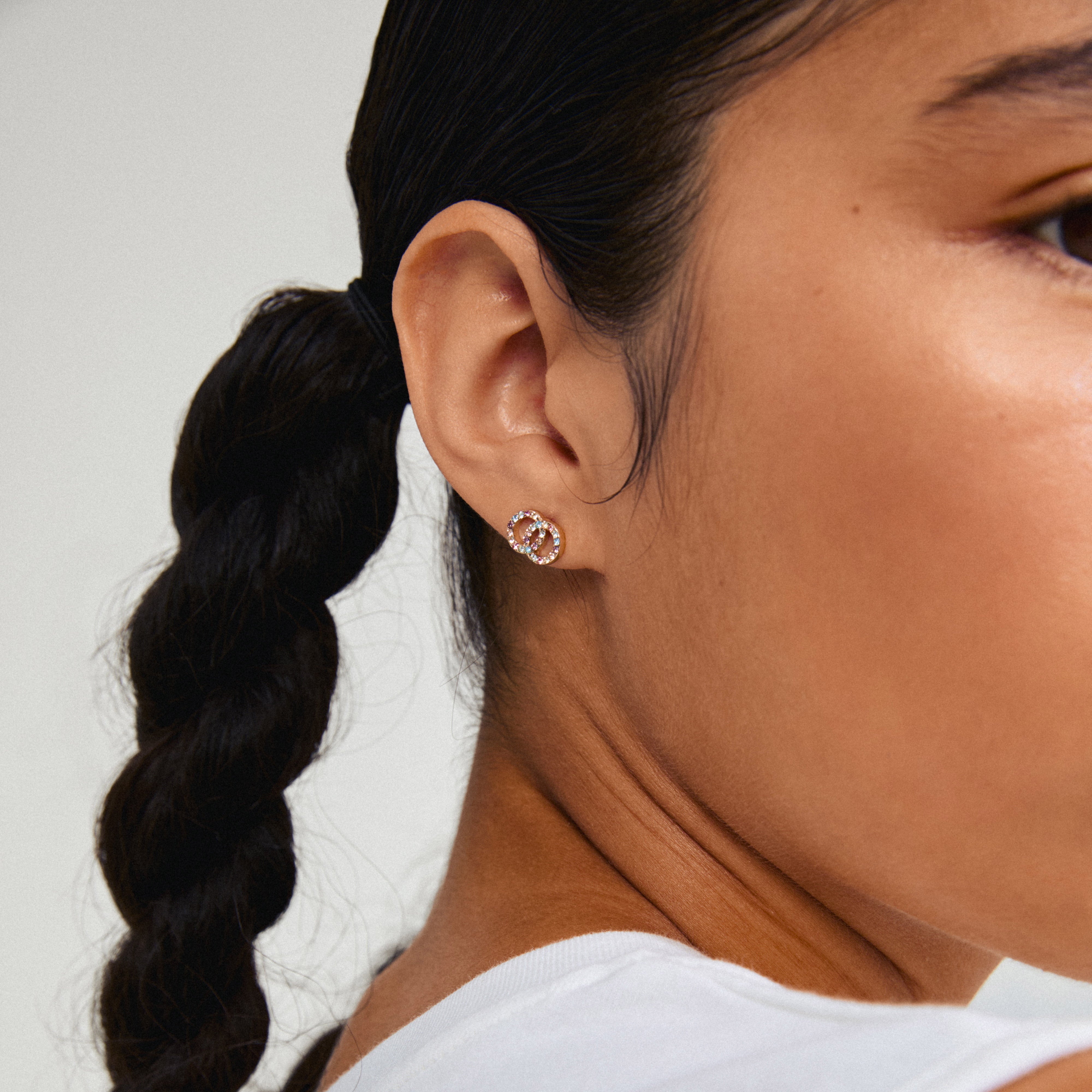 JOLINA recycled earrings, gold-plated with multi-colour crystals, adorn a woman's ear, showcasing intertwined halo design from Pilgrim.