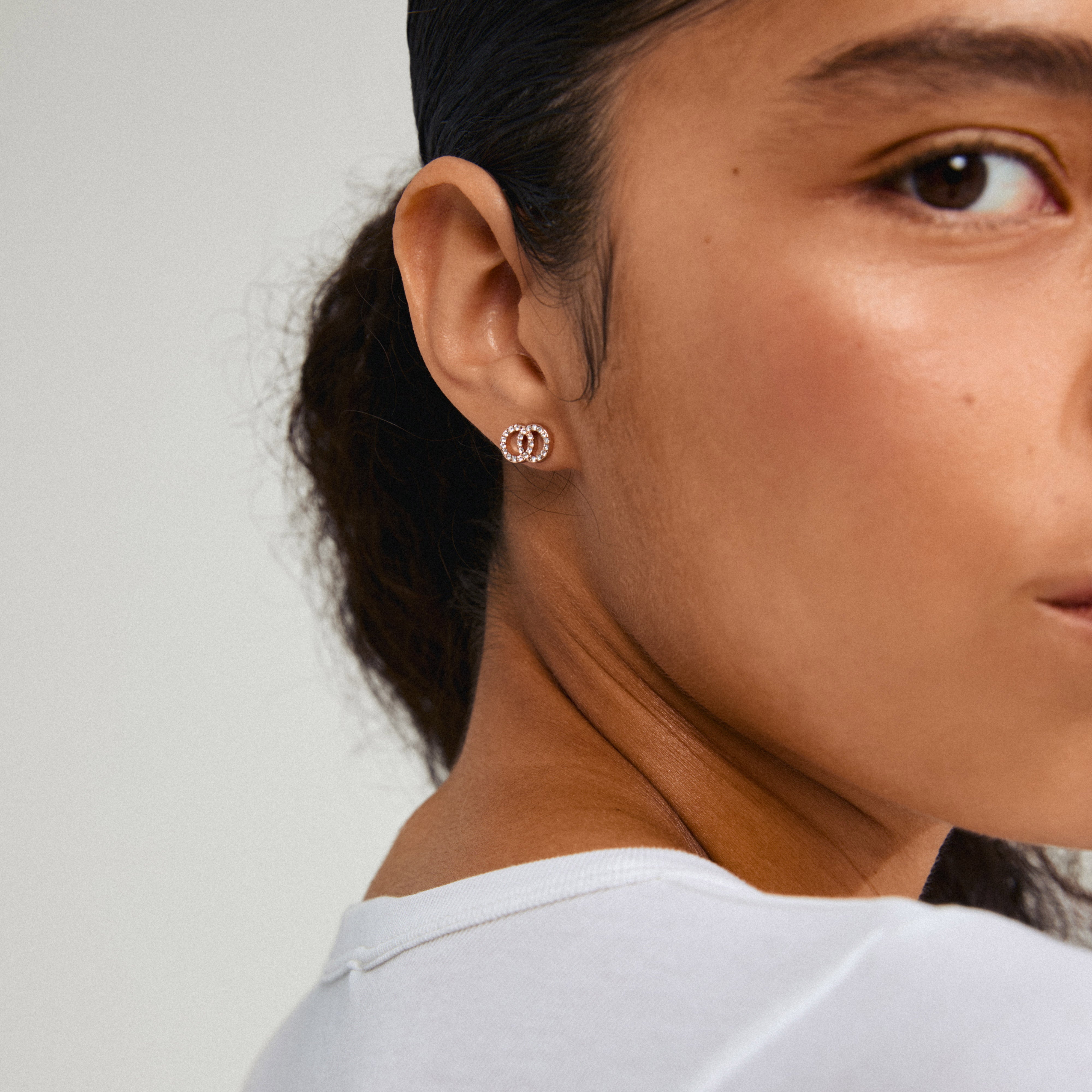 JOLINA recycled earrings in rose gold-plated design, featuring two intertwined halos with sparkling crystals, showcased on a woman's ear.