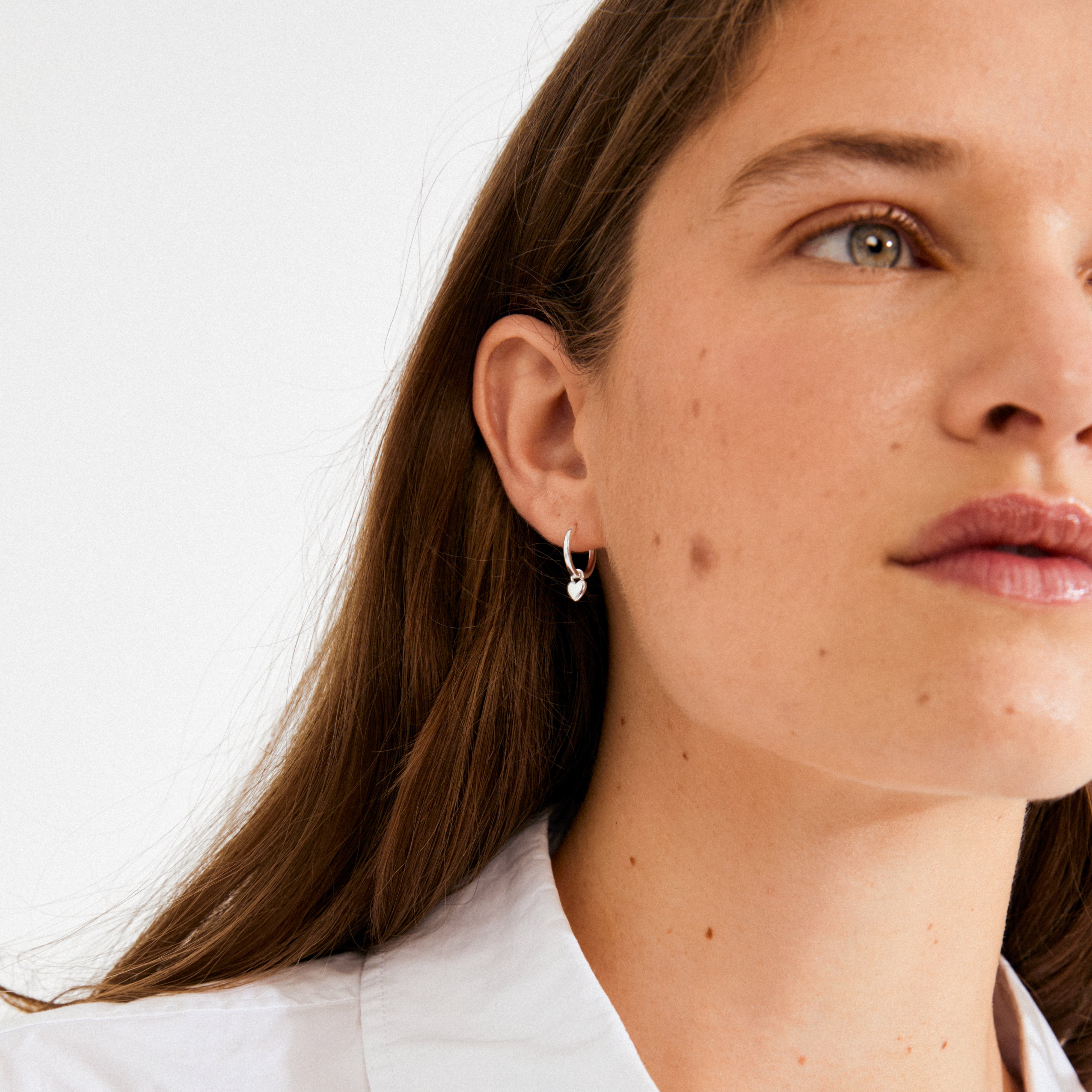 Young woman wearing ALAIA earrings silver-plated, featuring a small hoop with a dangling heart charm, showcasing Pilgrim's blend of timeless romance and modern style.