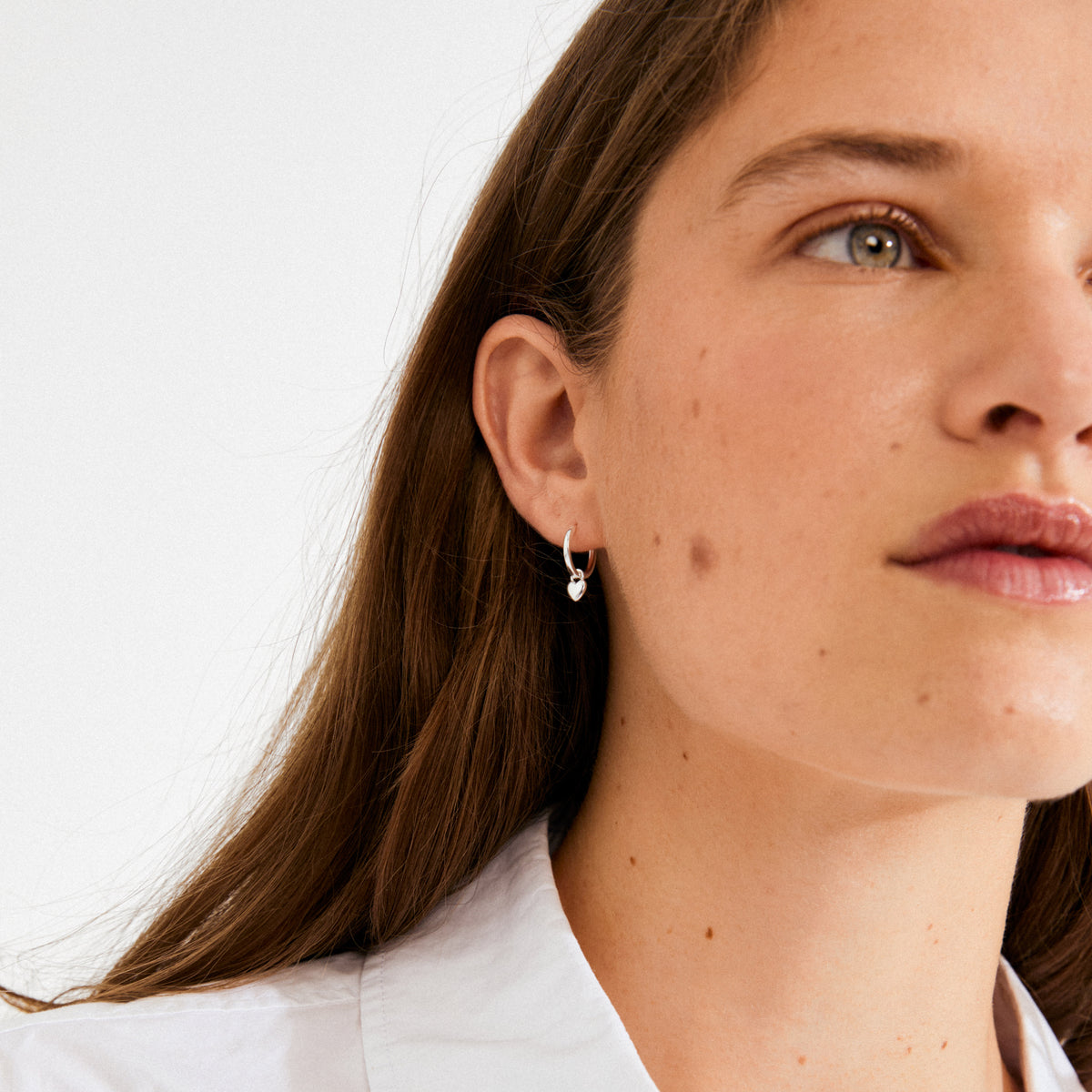 Young woman wearing ALAIA earrings, silver-plated huggie hoops with a heart pendant, showcasing a romantic, versatile design crafted from recycled materials for everyday or special occasions.