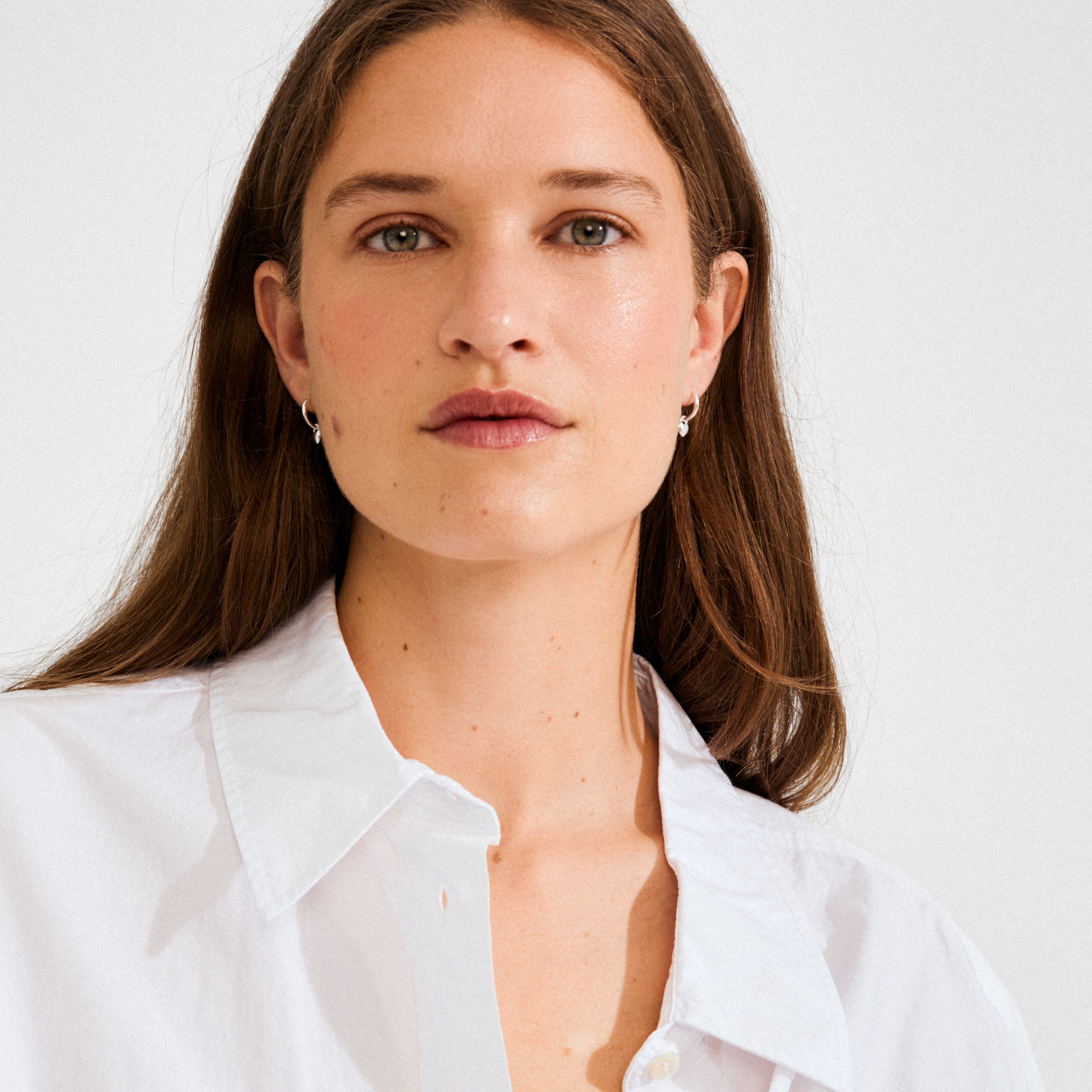 Young woman wearing ALAIA earrings—silver-plated huggie hoops with petite heart pendants, styled for a contemporary and romantic look. Perfect for versatile, everyday wear.