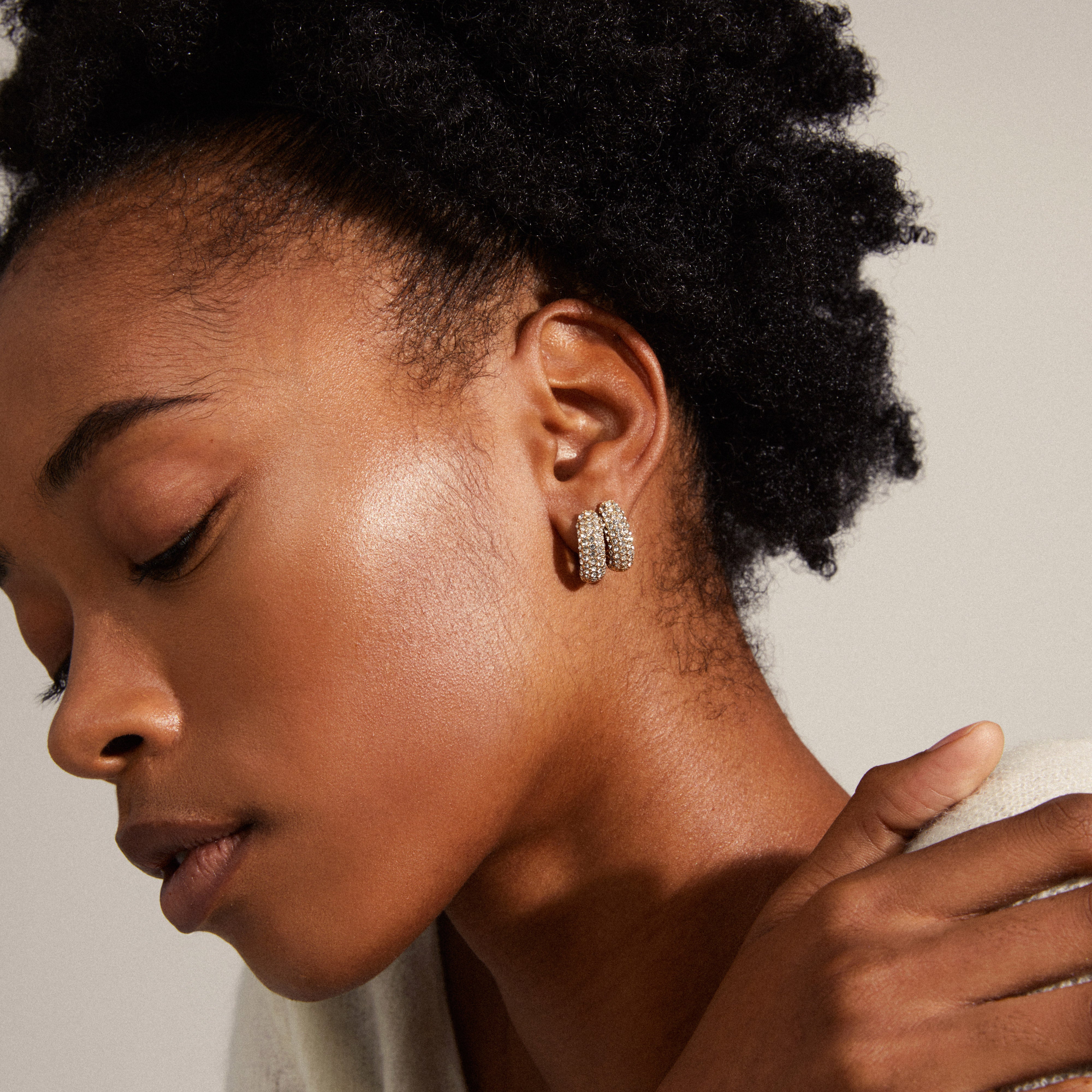 LONA chunky crystal huggie hoops, gold-plated, elegantly adorn a woman's ear, showcasing sparkling Preciosa crystals for a sophisticated, eco-friendly style.