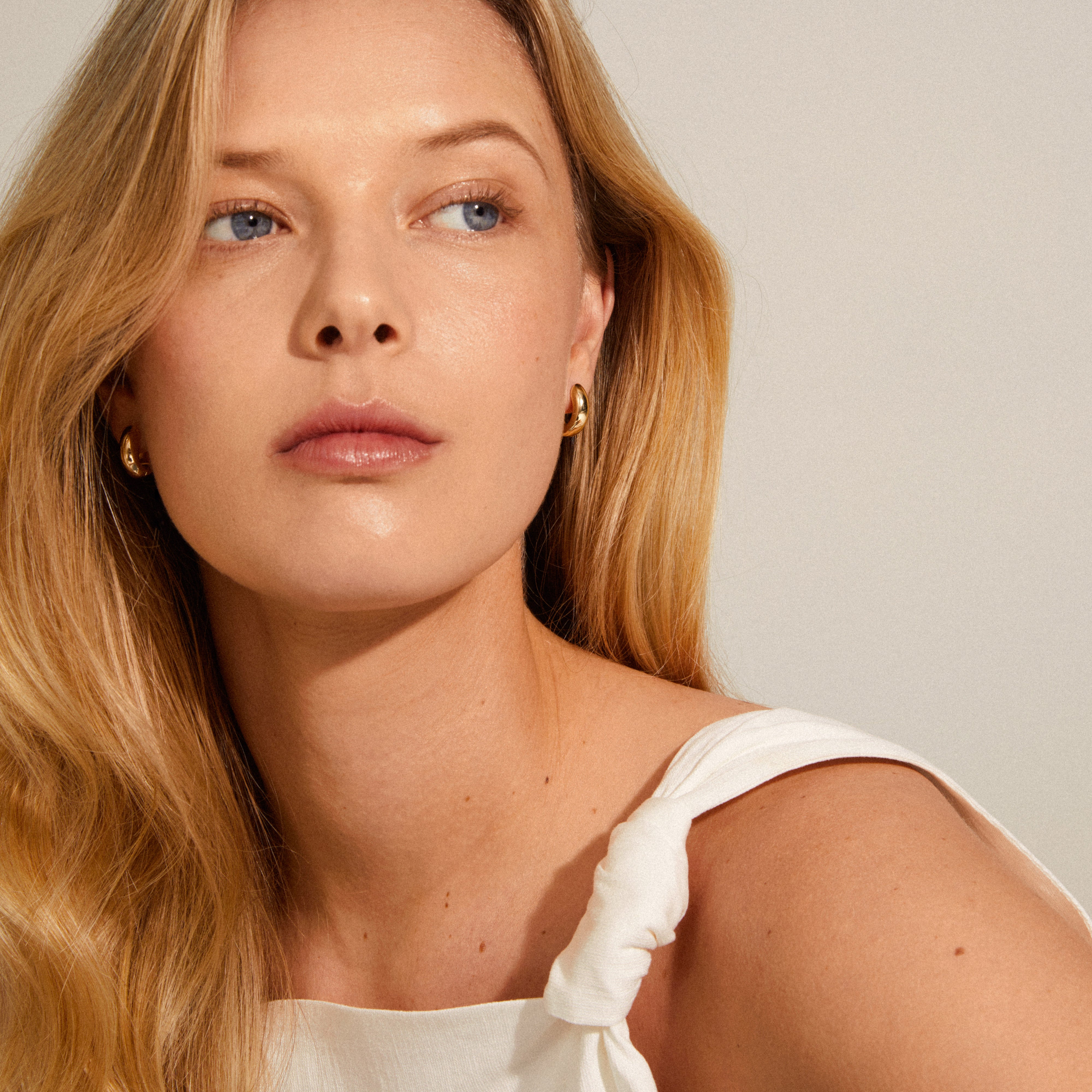 MINE chunky huggie hoops, gold-plated, adorn a woman looking away, highlighting their elegant, shiny design, perfect for a sophisticated, feminine style.