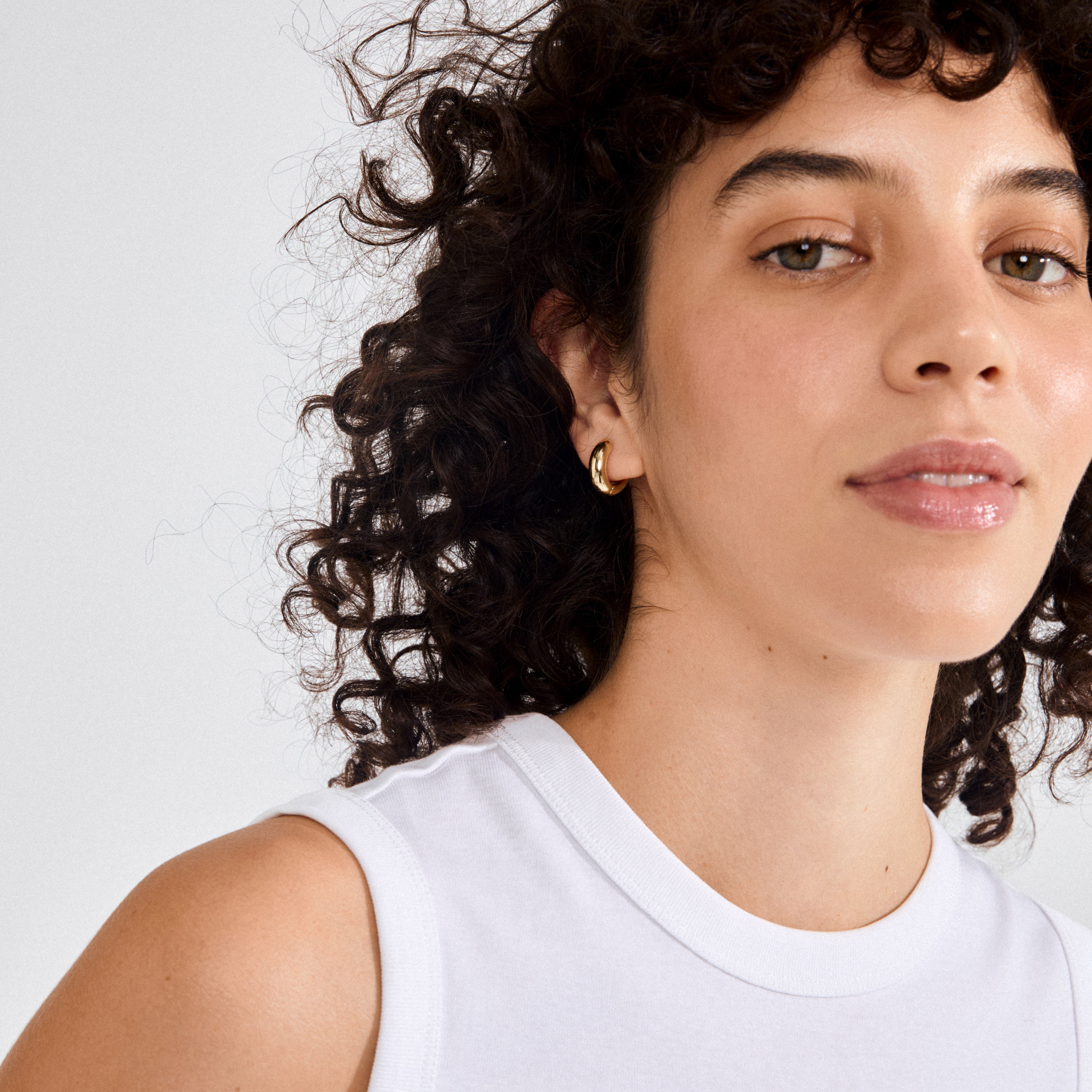 Young adult wearing MINE chunky huggie hoops gold-plated, showcasing their high-shine, sculpted design. Earrings complement a minimal-makeup look with a subtle, stylish flair.