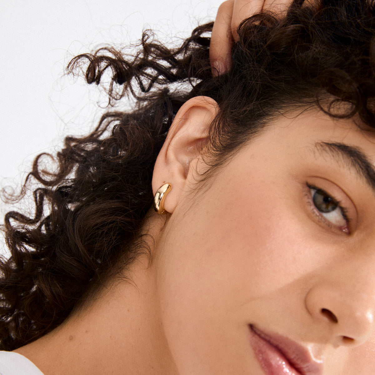 Close-up of MINE chunky huggie hoops gold-plated earring on an ear, with curly hair and smooth skin, showcasing Pilgrim's elegant and sustainable design.