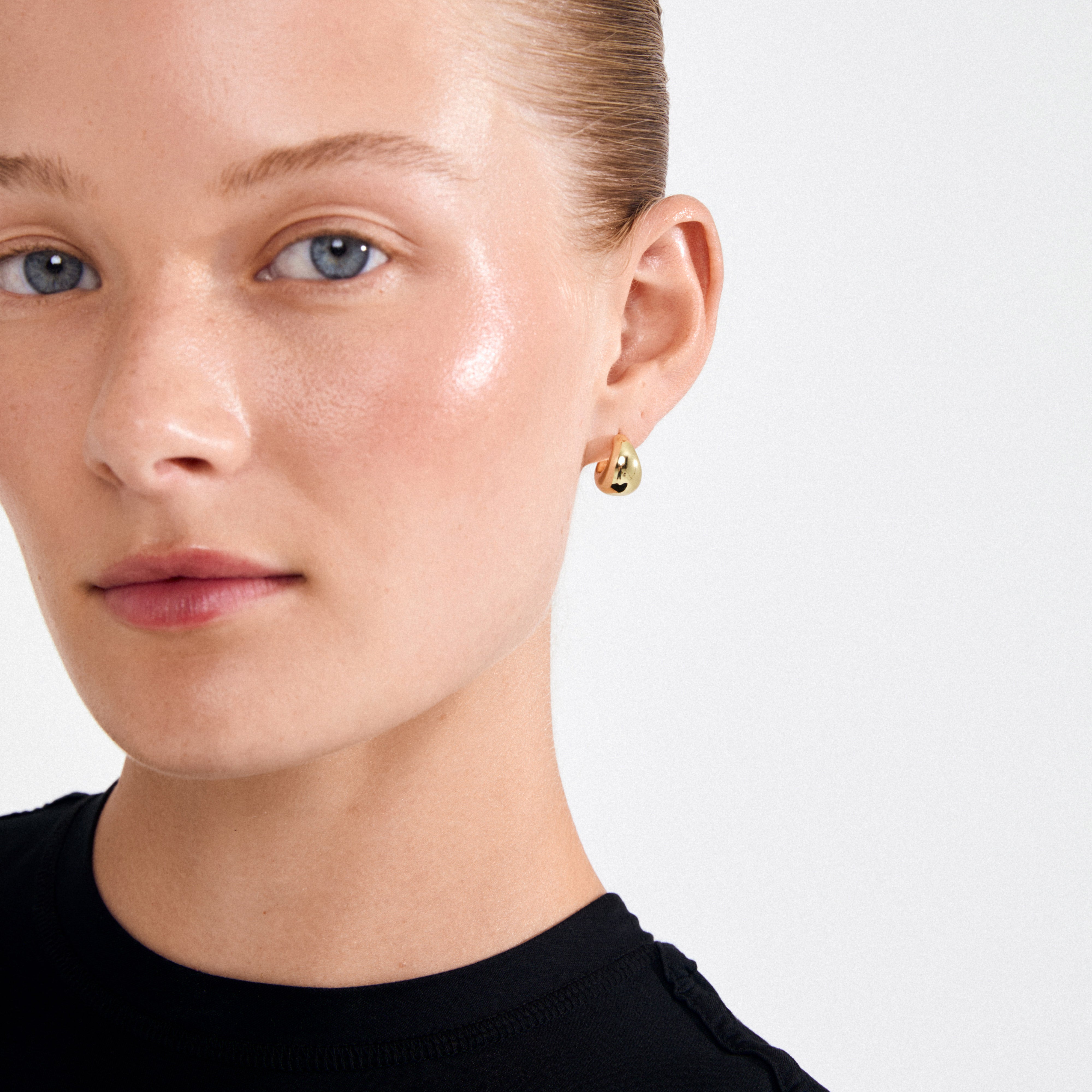 Young woman wearing a black top showcases ALEXANE gold-plated teardrop earrings, highlighting their glossy, retro design against slicked-back hair and a natural look.