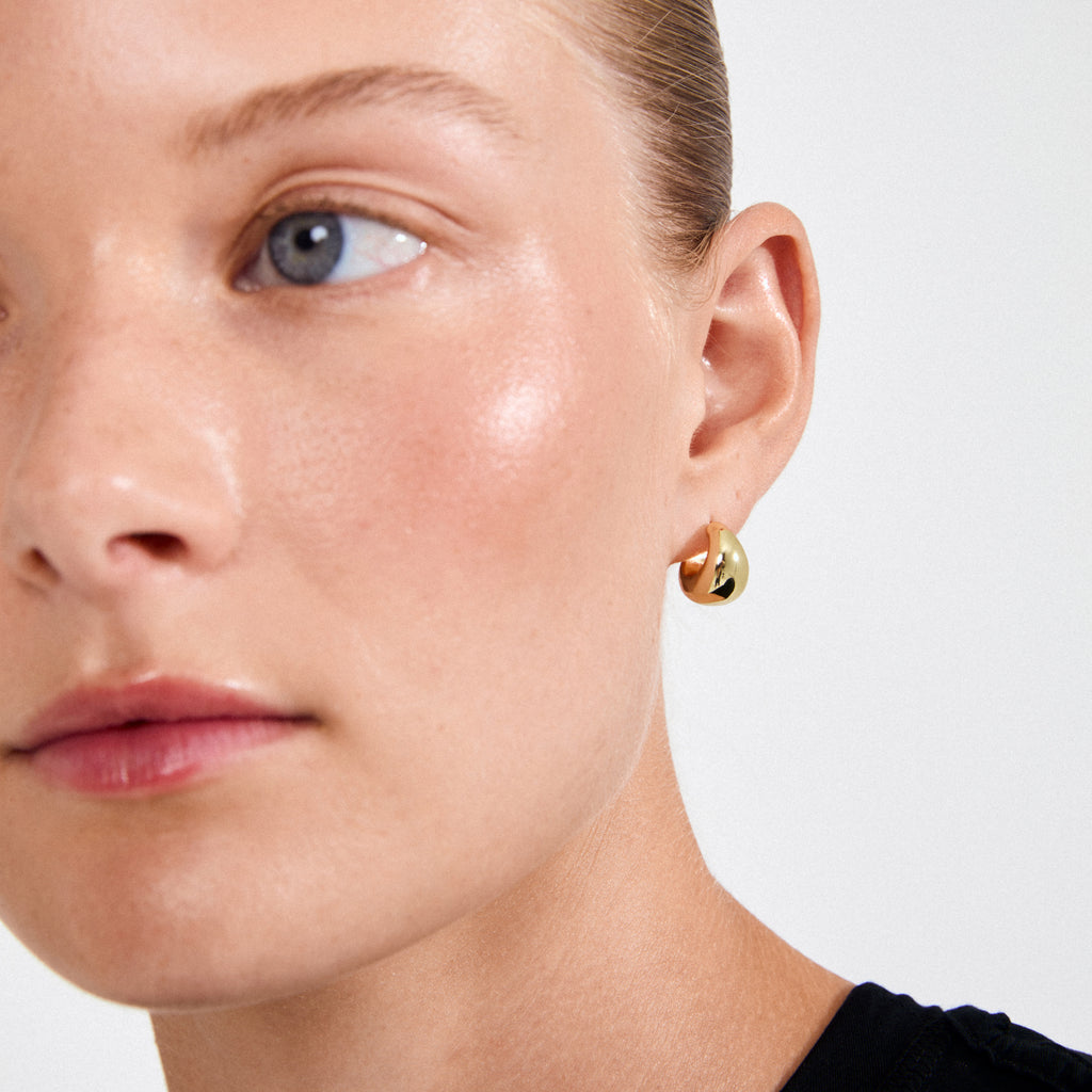 Young woman wearing ALEXANE chunky mini hoop earrings, gold-plated, showcasing their glossy droplet shape and smooth, retro-inspired curves against her understated look.