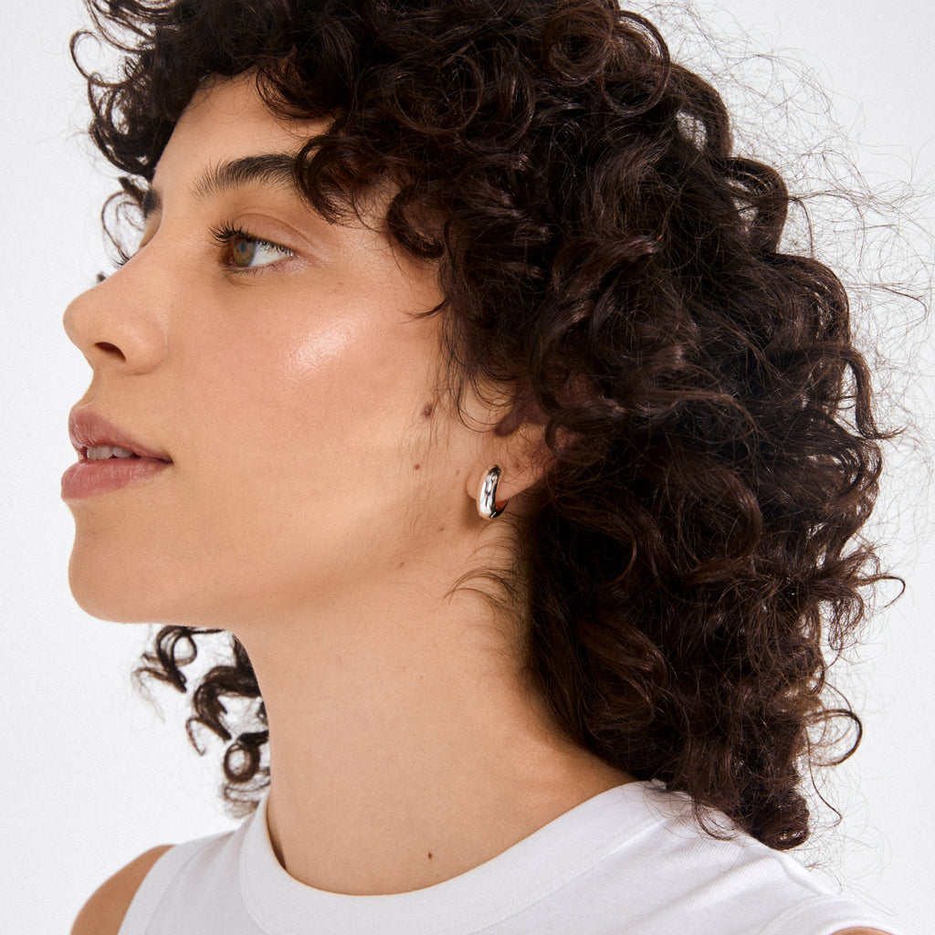 Young person wearing MINE chunky huggie hoops, silver-plated, showcasing their polished, high-shine finish and softly sculpted design against a plain white background.