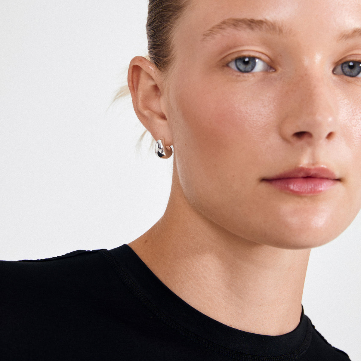 Young woman wearing ALEXANE chunky mini hoop earrings, silver-plated with a glossy, droplet-inspired design, showcasing a subtle retro cool and timeless elegance.