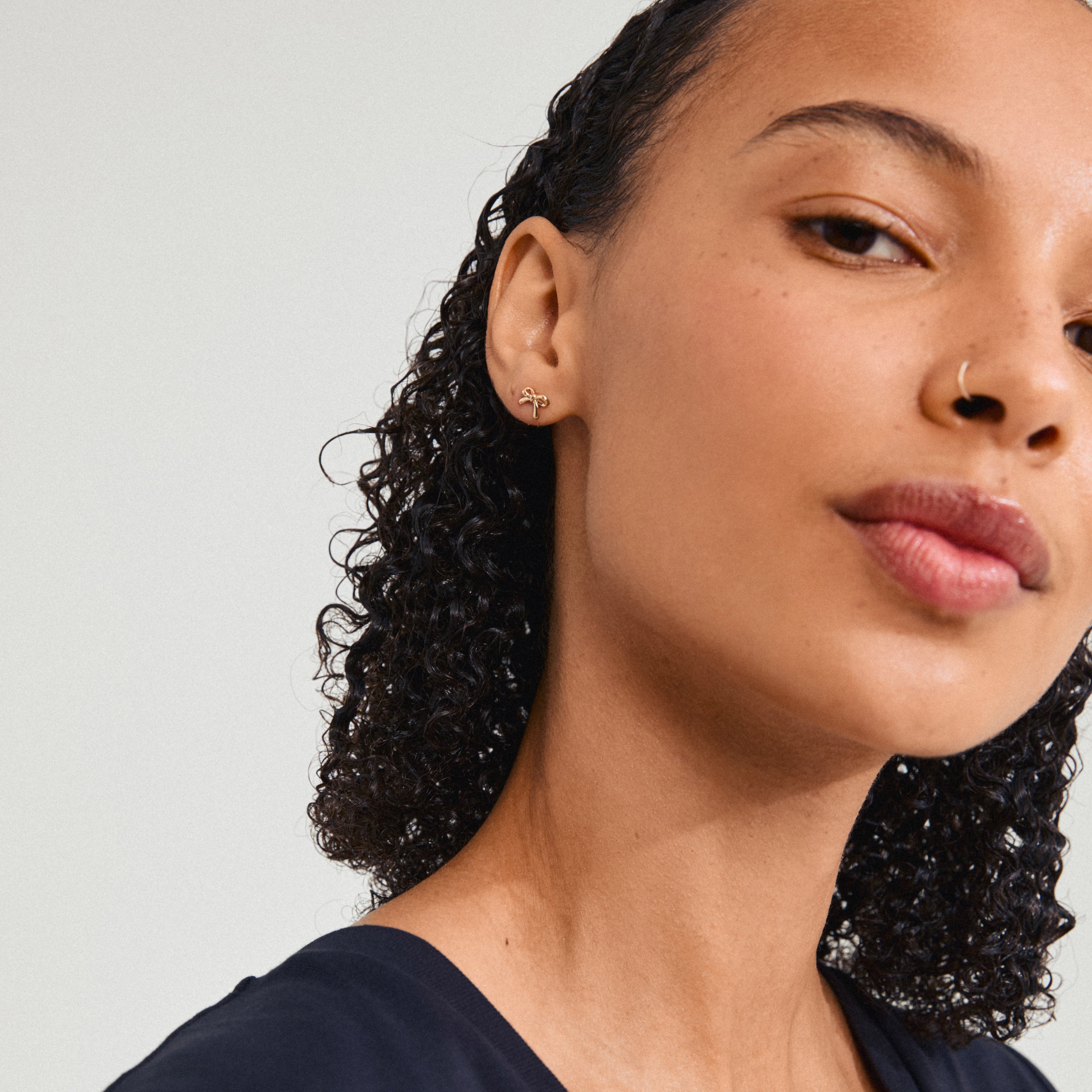 LYSANDRA earrings gold-plated, worn by a woman with curly hair and a nose ring, emphasize a trendy bow design, perfect for stylish, everyday looks.