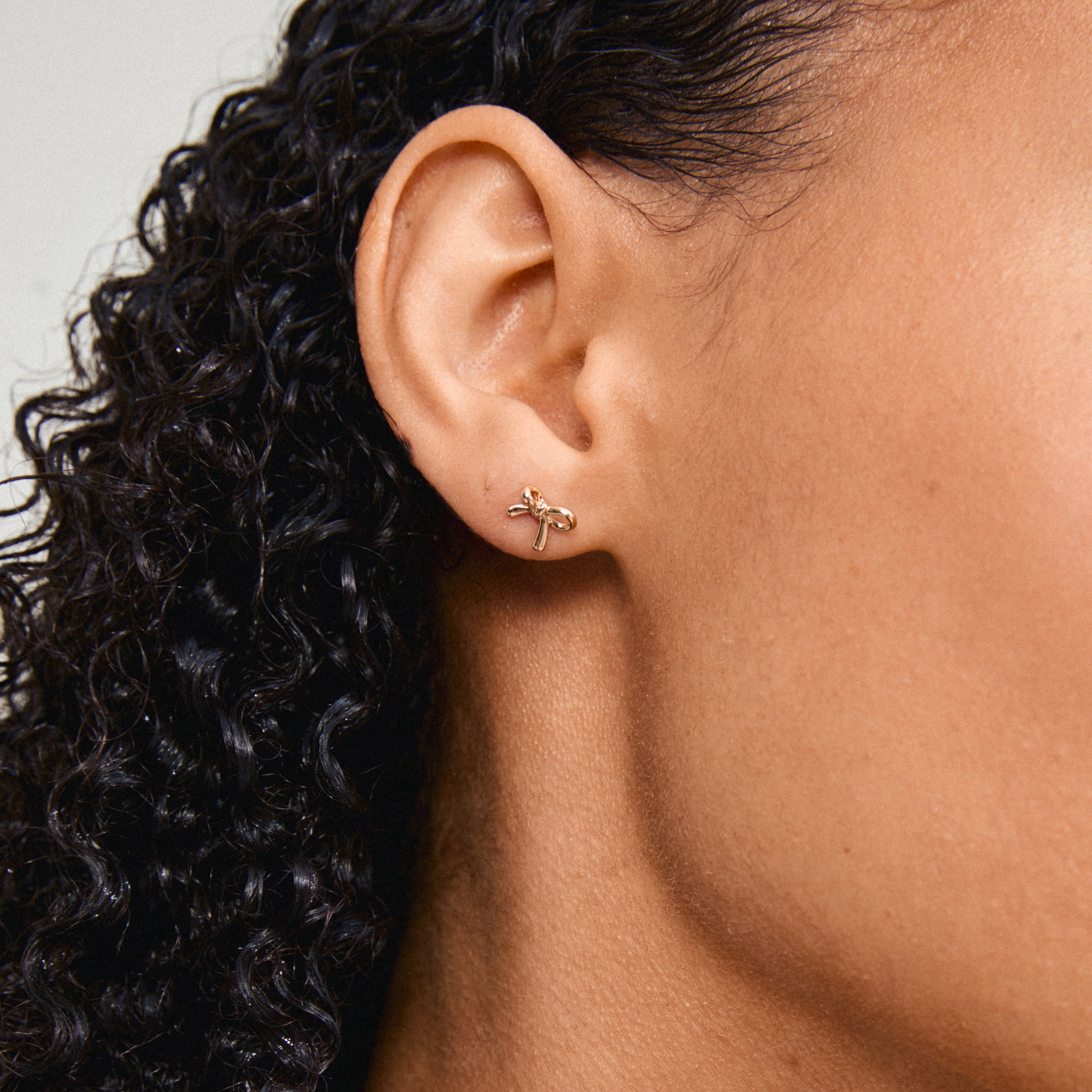 Close-up of a woman's ear wearing LYSANDRA gold-plated bow earrings, highlighting the trendy, sustainable design made from 97% recycled materials.