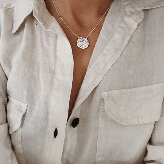 MARLEY necklace silver-plated with a textured medallion, inspired by ancient amulets, elegantly hangs on a model wearing a light linen shirt, showcasing its rustic charm.