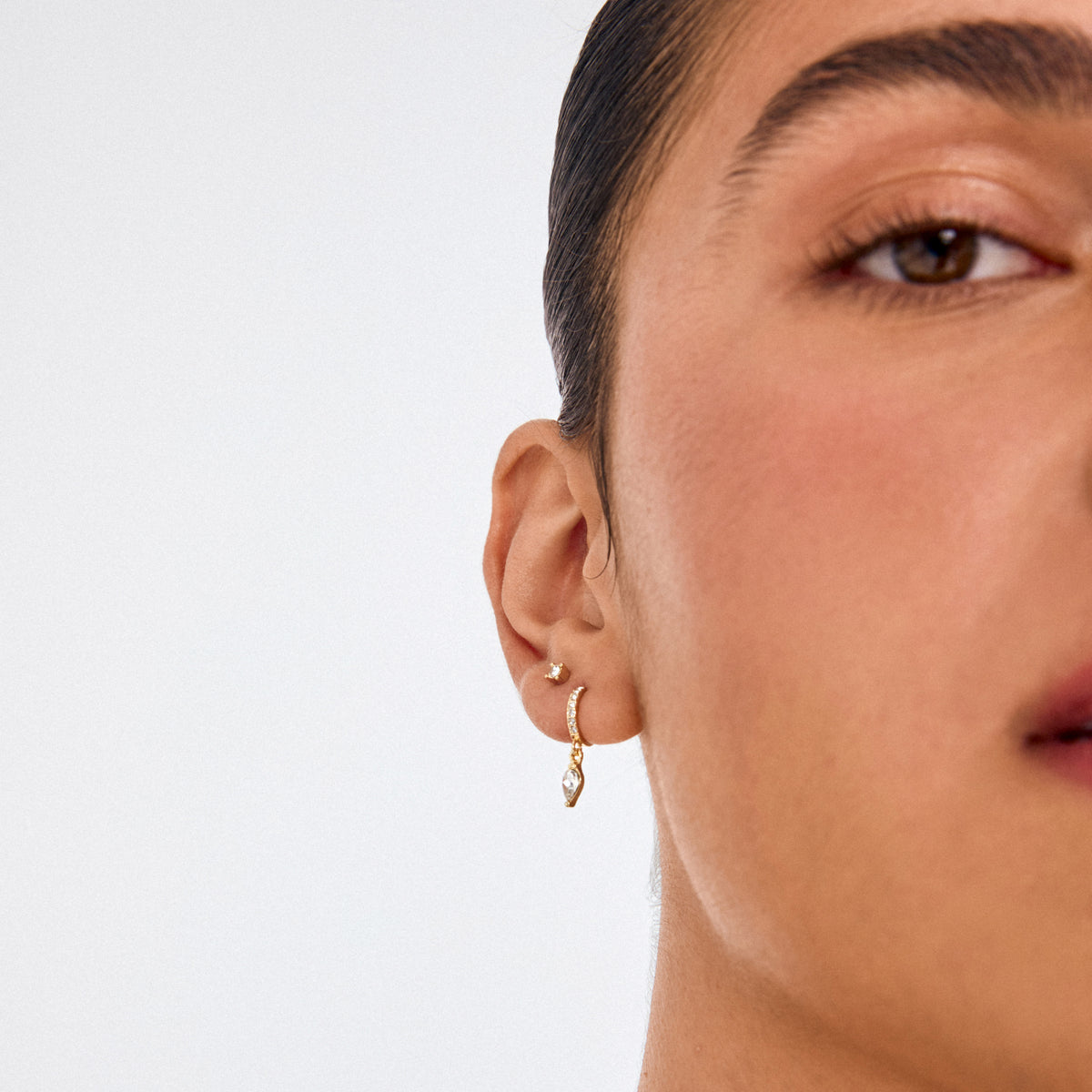 ELZA crystal earrings 2-in-1 set gold-plated featuring a stud and gemstone-studded hoop with teardrop pendant, worn on a person’s ear.
