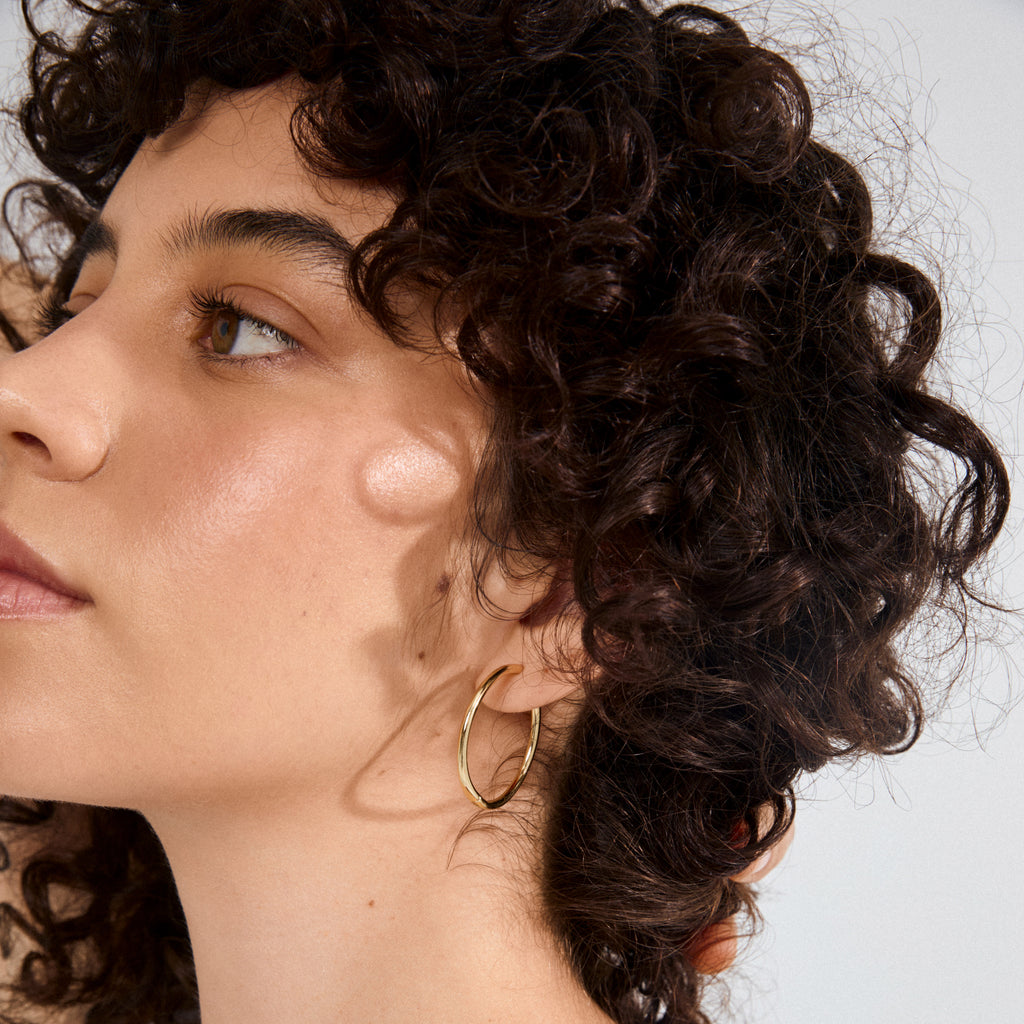 Side-profile of a person wearing Pilgrim's EANNA maxi hoops gold-plated, showcasing the simple gold hoop earring against their ear with soft lighting.