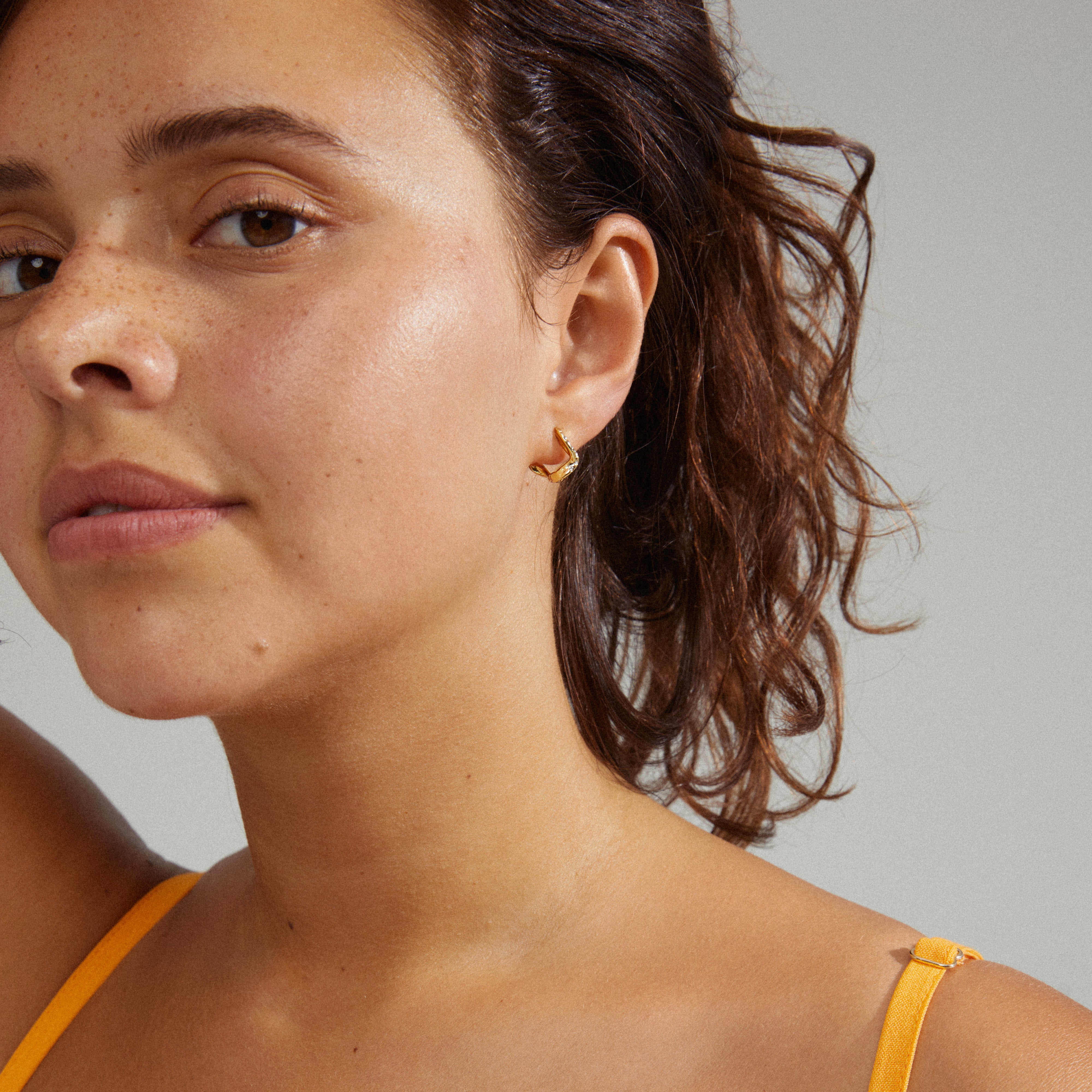 EVRA organic shaped crystal hoops gold-plated, featuring a curvy design with shiny Preciosa crystals and easy click lock, worn by a woman with brown hair.
