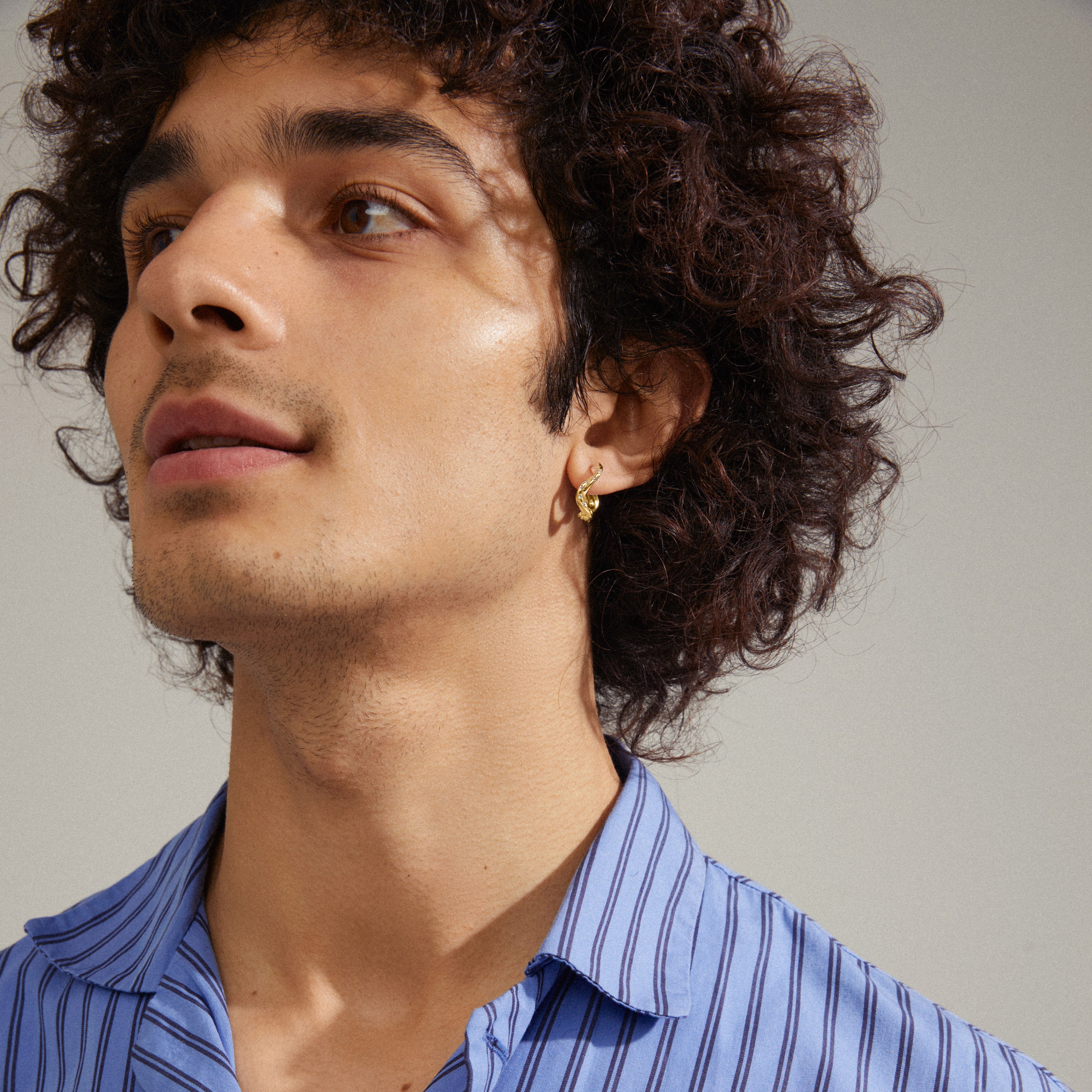 Man wearing blue striped shirt showcasing Evra organic shaped gold-plated crystal hoops. The earrings feature a wavy design and discreet shine, highlighting their elegant craftsmanship.