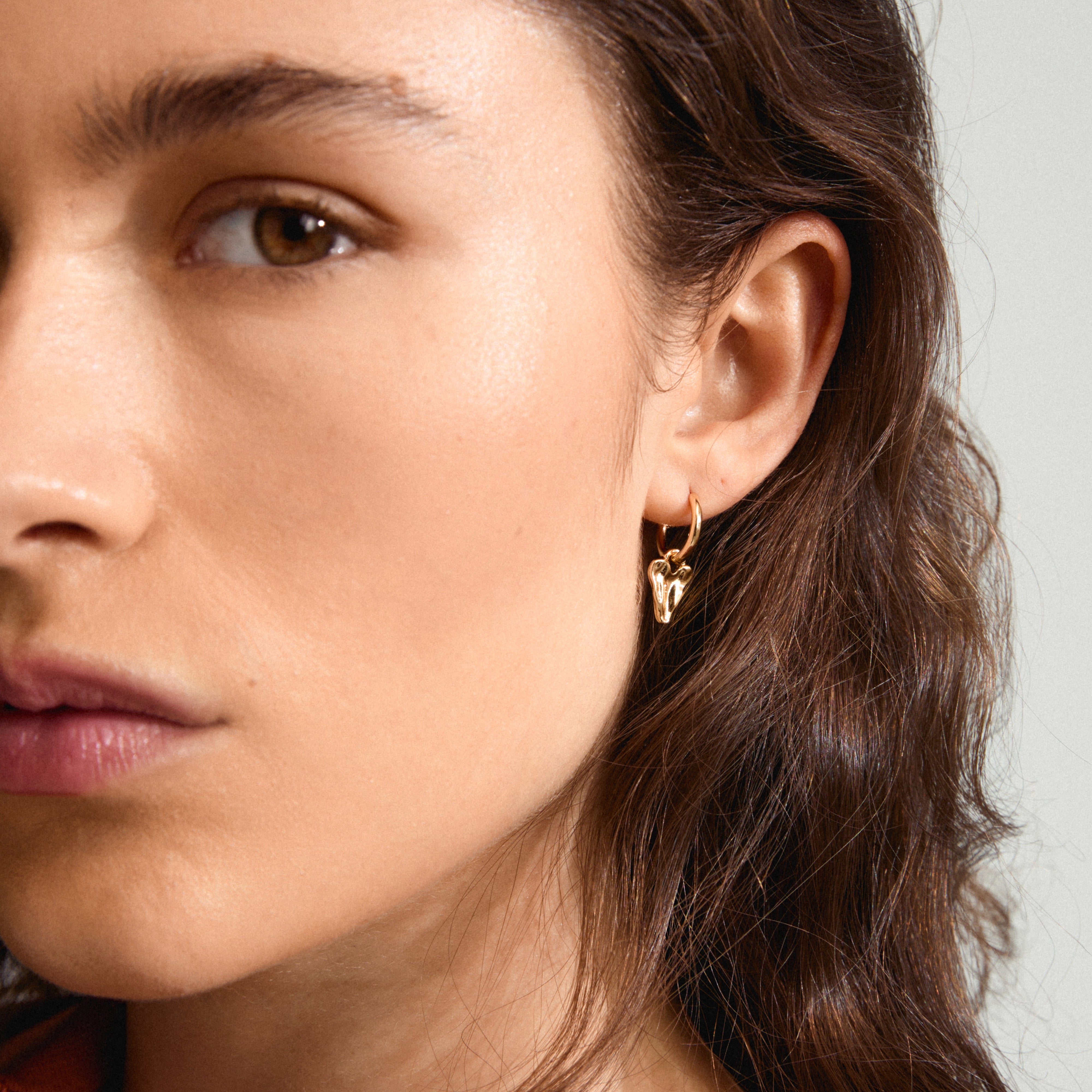 Close-up of a woman's face showcasing JULES gold-plated earrings, featuring a small heart pendant, highlighting Scandinavian craftsmanship and modern design.