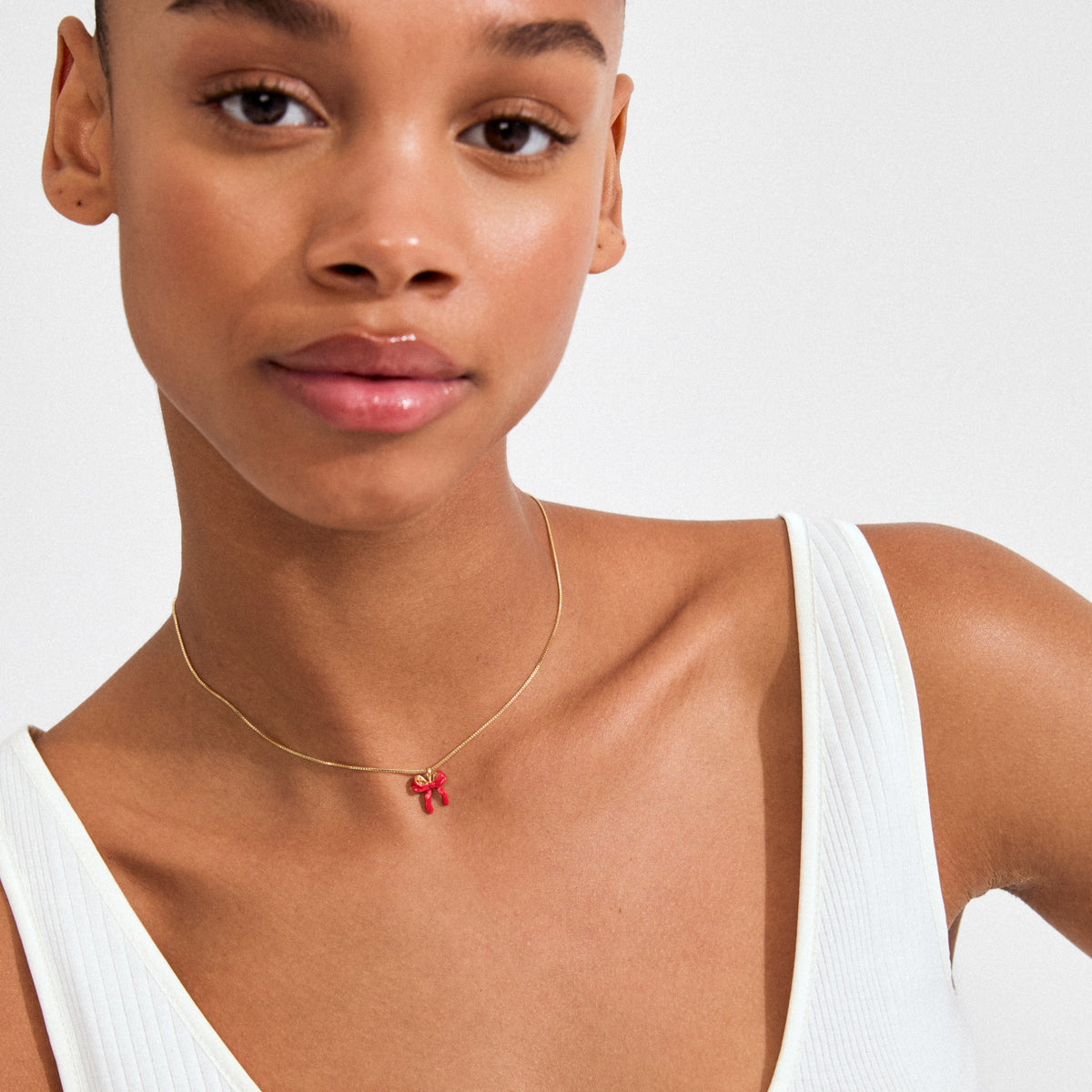 Young woman wearing Pilgrim's SIGRUN necklace, featuring a gold-plated chain with a small red enamel bow pendant, styled with a white ribbed tank top.