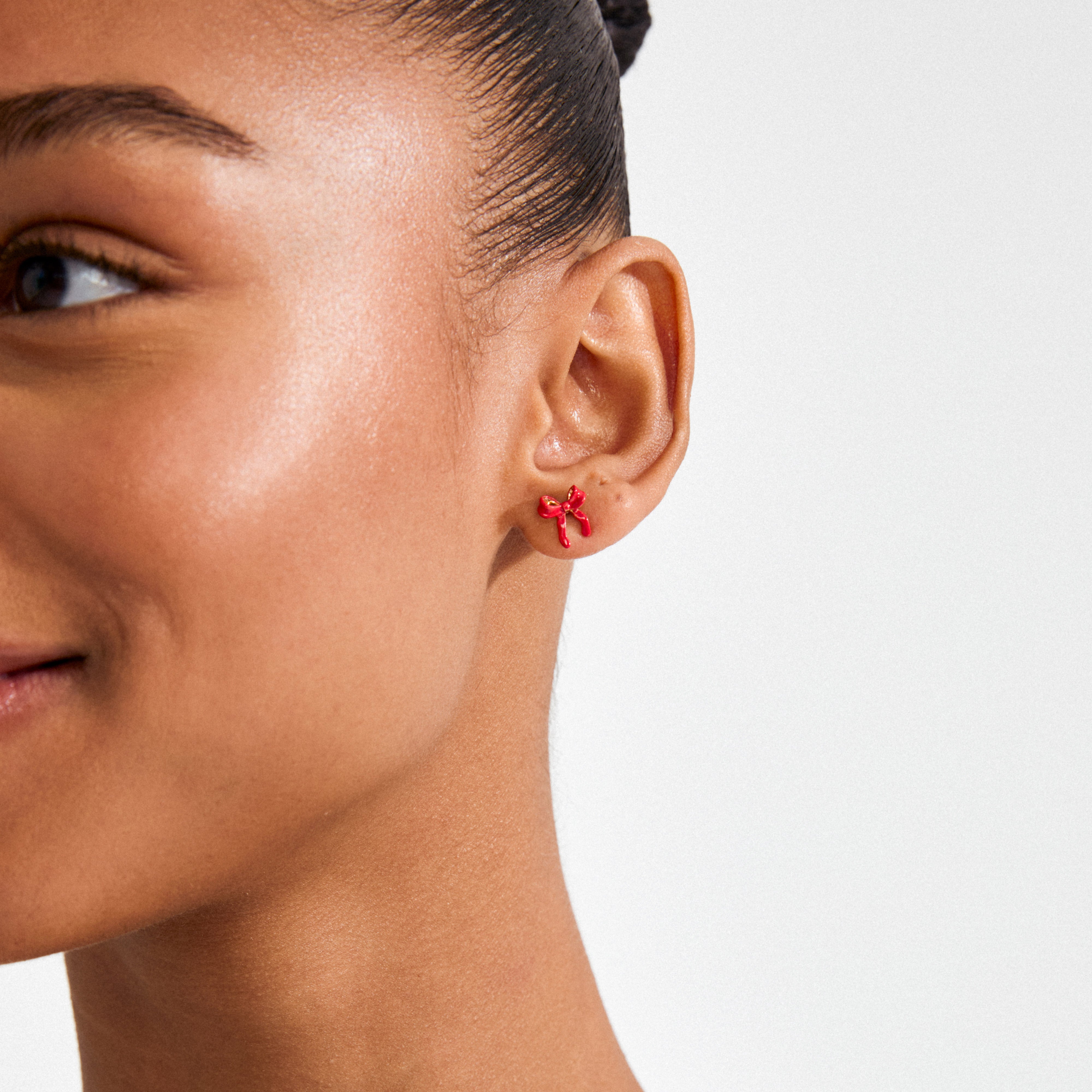 Close-up of a person wearing SIGRUN gold-plated earrings by Pilgrim, featuring small red enamel bows, perfect for adding a trendy touch to minimalist styles.