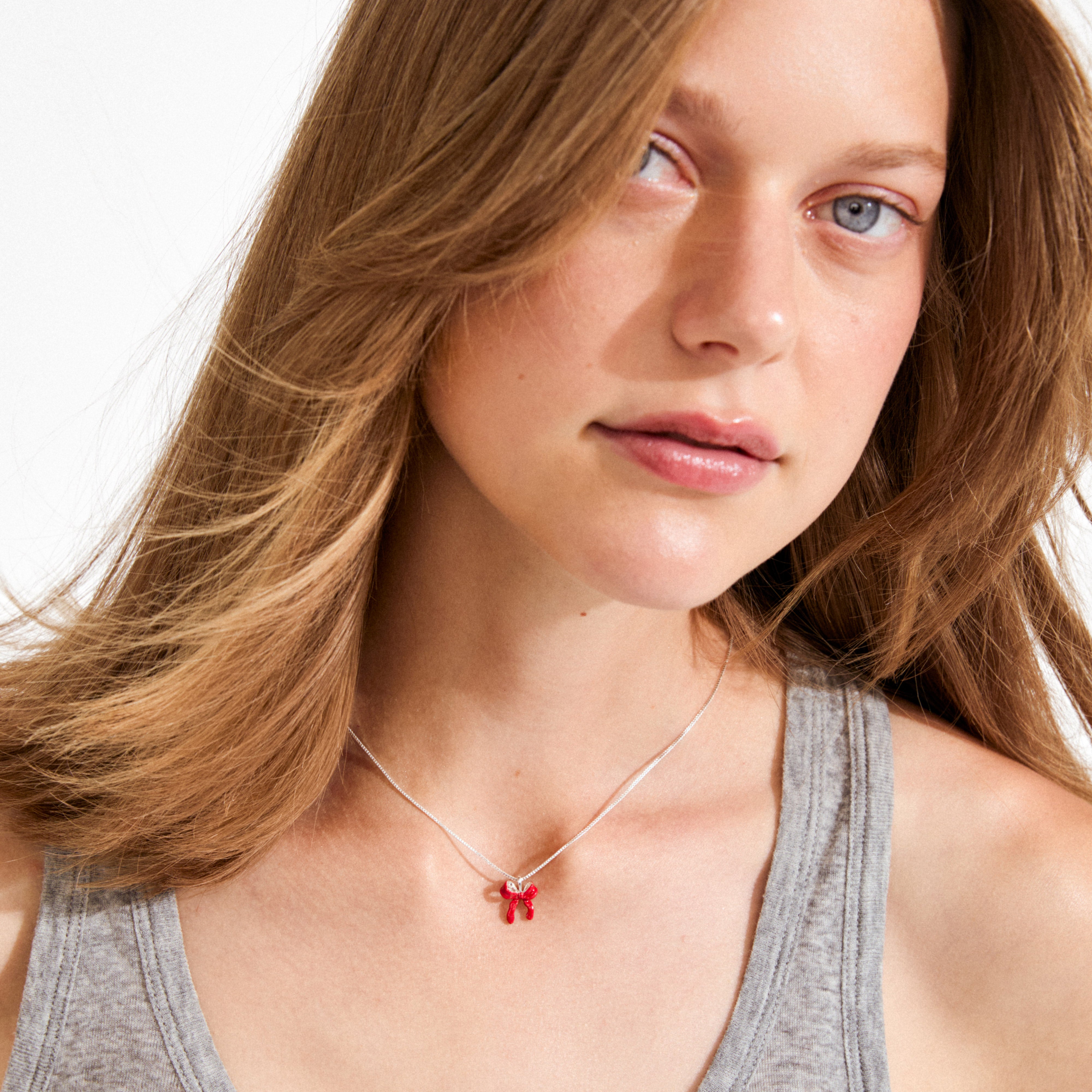 Young woman wearing the SIGRUN necklace, a silver-plated piece from Pilgrim, featuring a small red enamel bow charm on a fine chain.