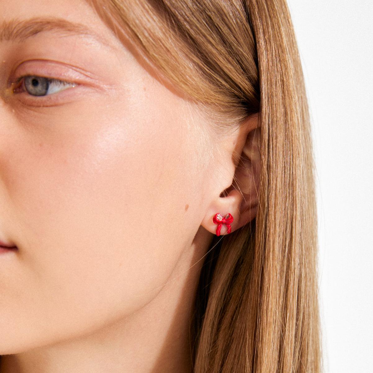 Close-up of a side view showing SIGRUN earrings, silver-plated, featuring a bright red bow-shaped stud, tucked behind light-brown/blonde hair.