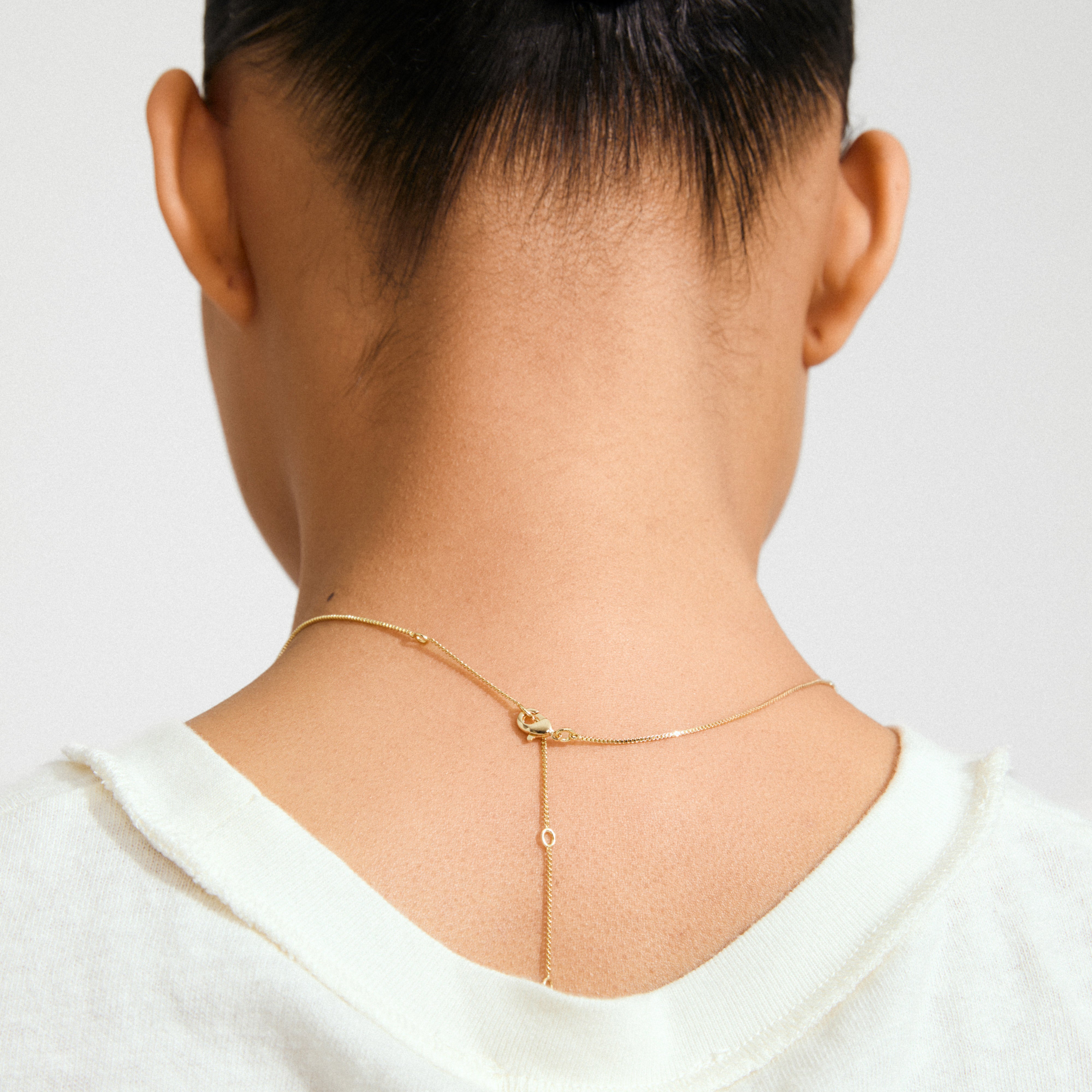 Person wearing the DAGMAR pendant necklace gold-plated with a delicate drop-chain detail, elegantly draped on the back of the neck over a white top.