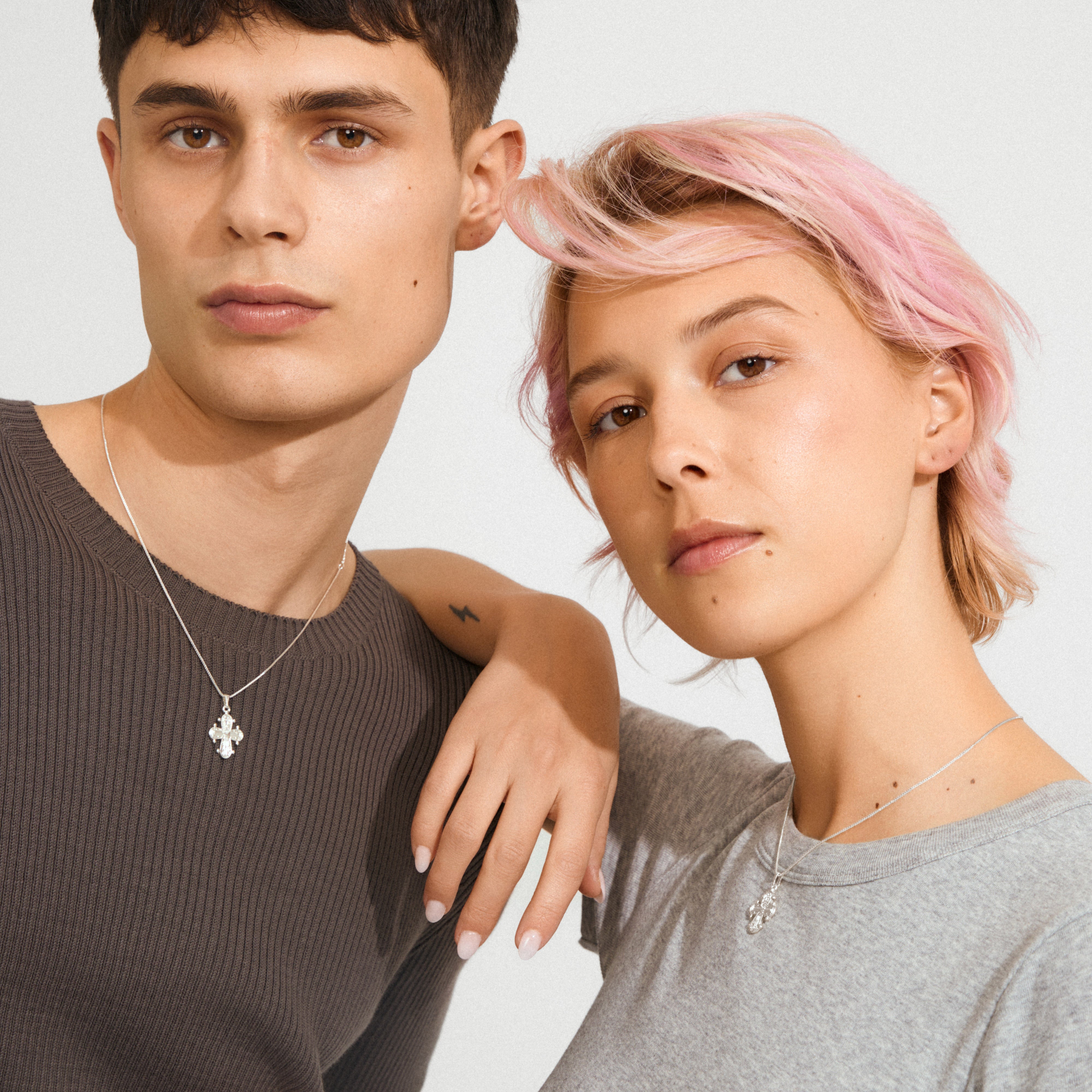 Two young adults showcase Pilgrim's DAGMAR pendant necklace, featuring silver-plated floral-style cross pendants on delicate chains, epitomizing modern Scandinavian elegance.