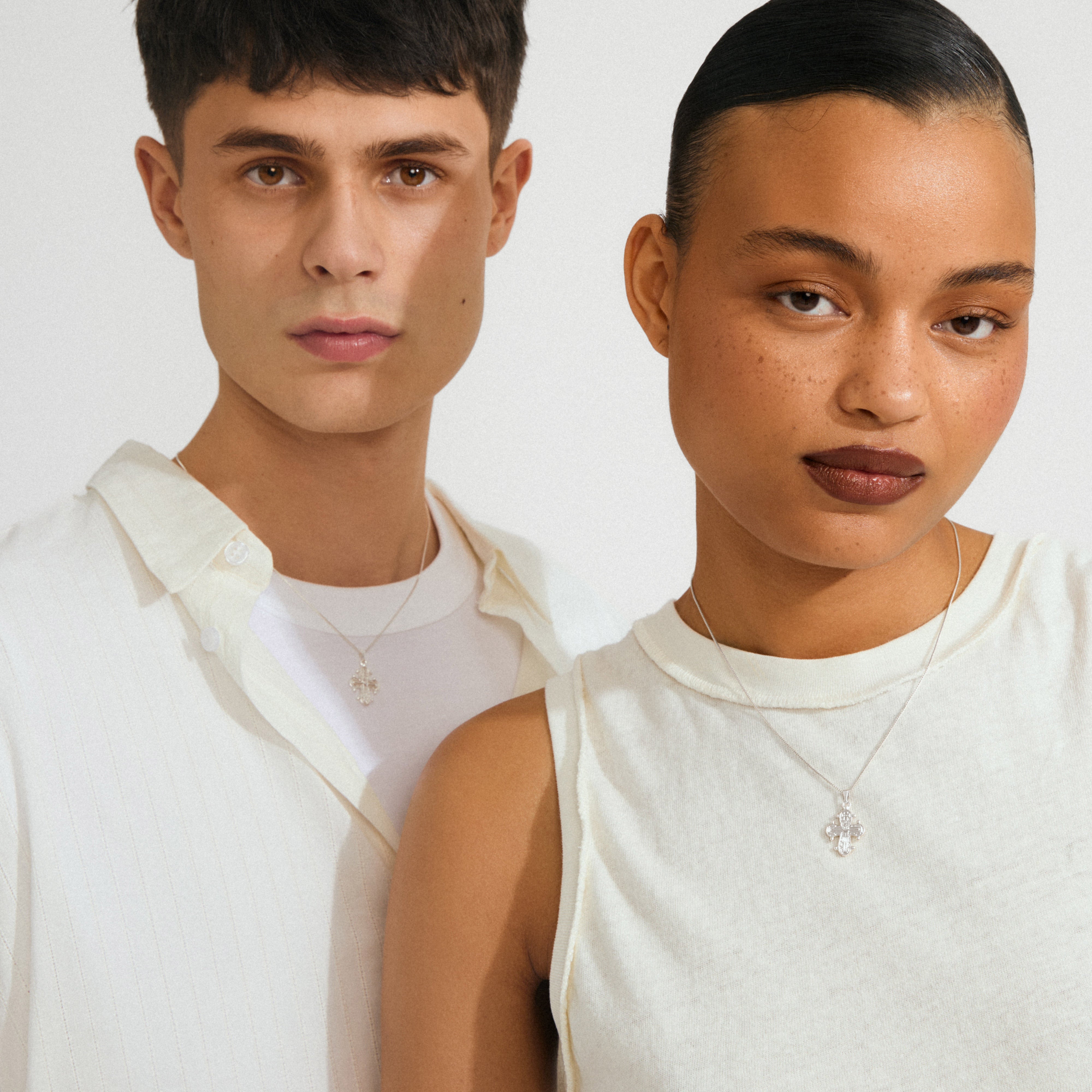 Two young adults wearing off-white tops and DAGMAR pendant necklace silver-plated, featuring small cross pendants, against a light background, embodying timeless Danish design.