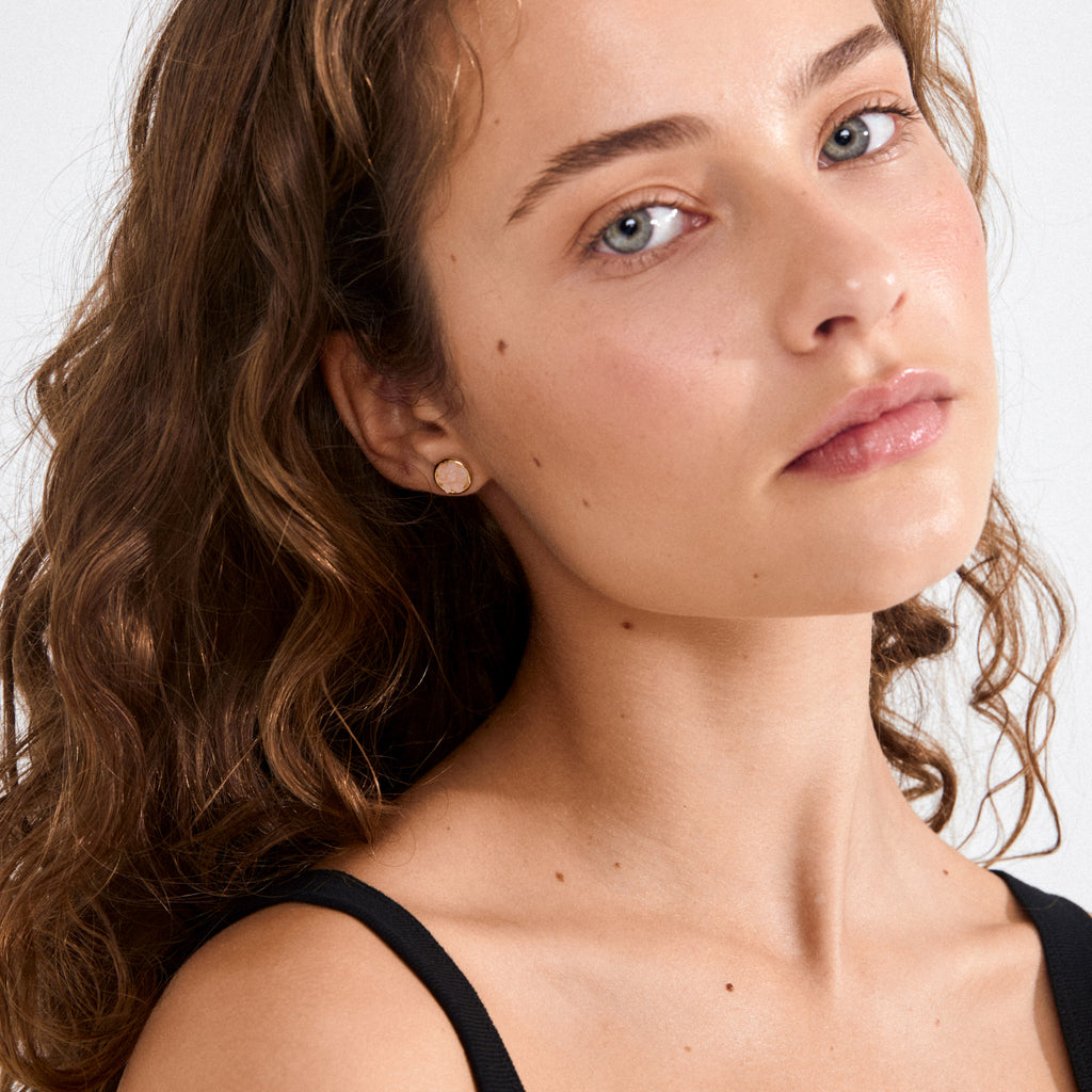 JOSEFINE earrings gold-plated with rustic pink stone in a textured, shiny setting, worn by a woman with wavy brown hair, showcasing edgy elegance.