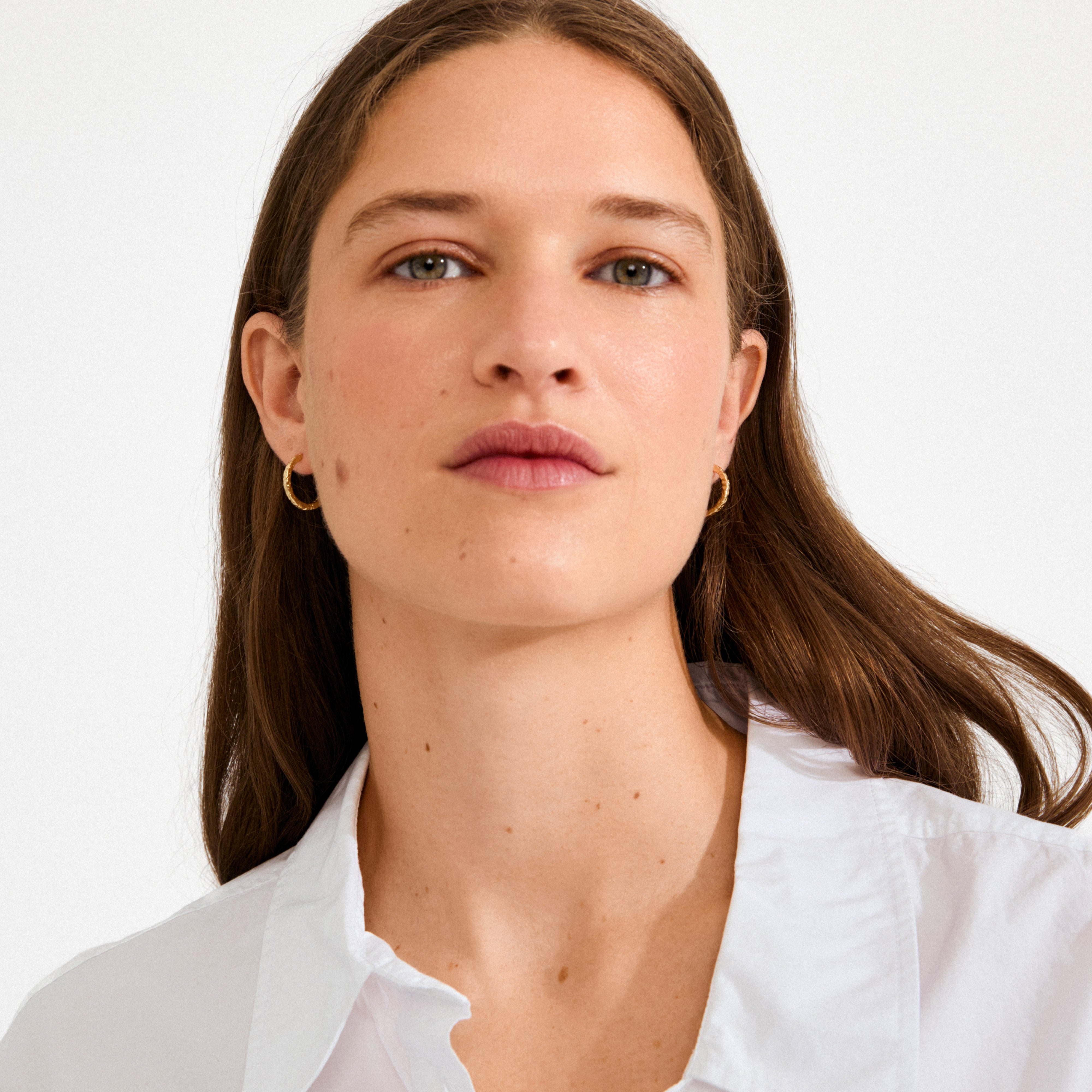Young woman wearing ELANOR rustic texture hoop earrings, gold-plated, showcasing their shiny finish and organic, nature-inspired design against a simple white shirt.