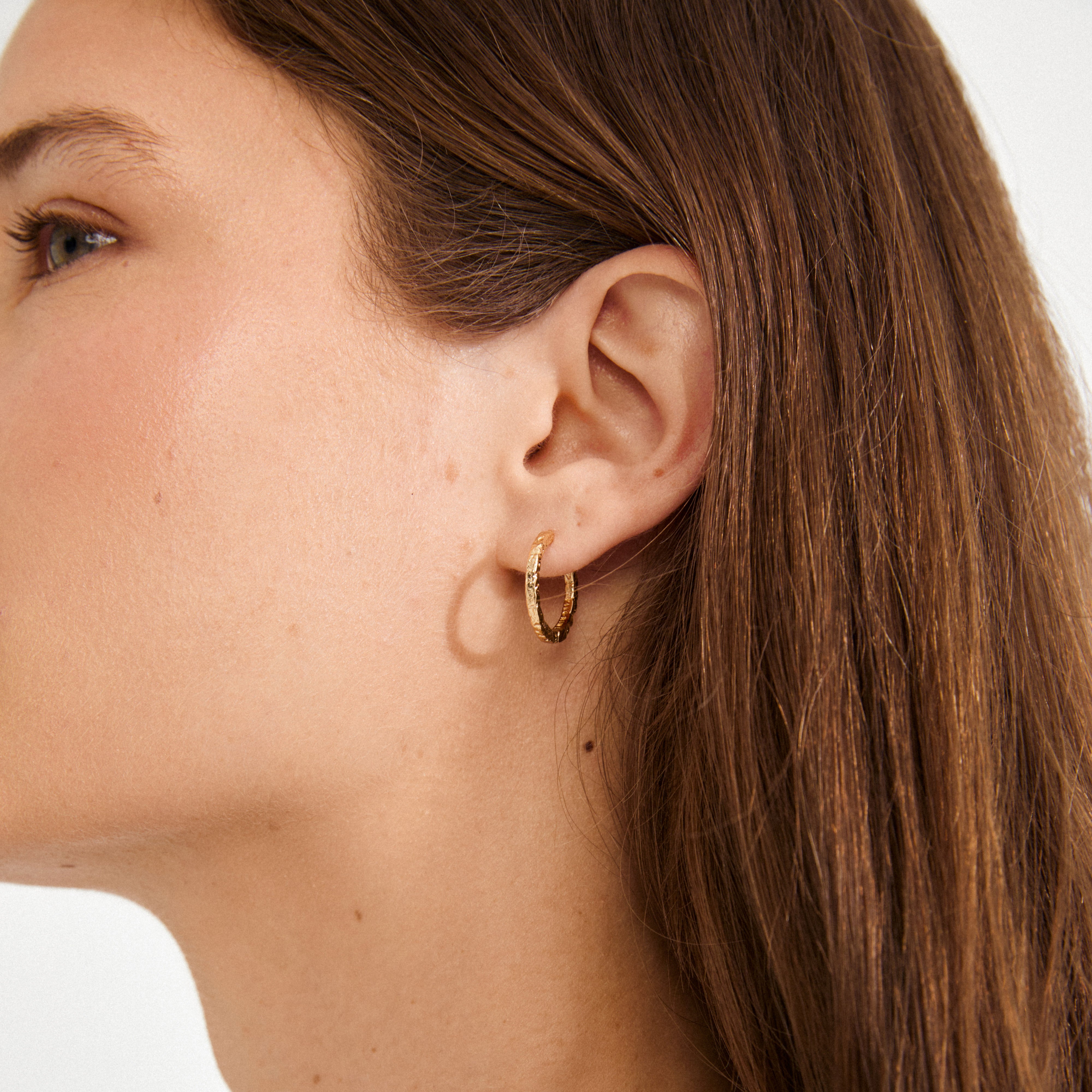 Close-up of a person wearing ELANOR rustic texture gold-plated hoop earrings by Pilgrim, featuring one textured and one smooth small hoop against a plain background.