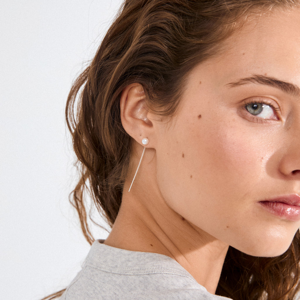 Profile view of a woman wearing Pilgrim's ELBA long chain pearl earrings, featuring a classic pearl stud with a silver-plated chain elegantly draping behind the ear.