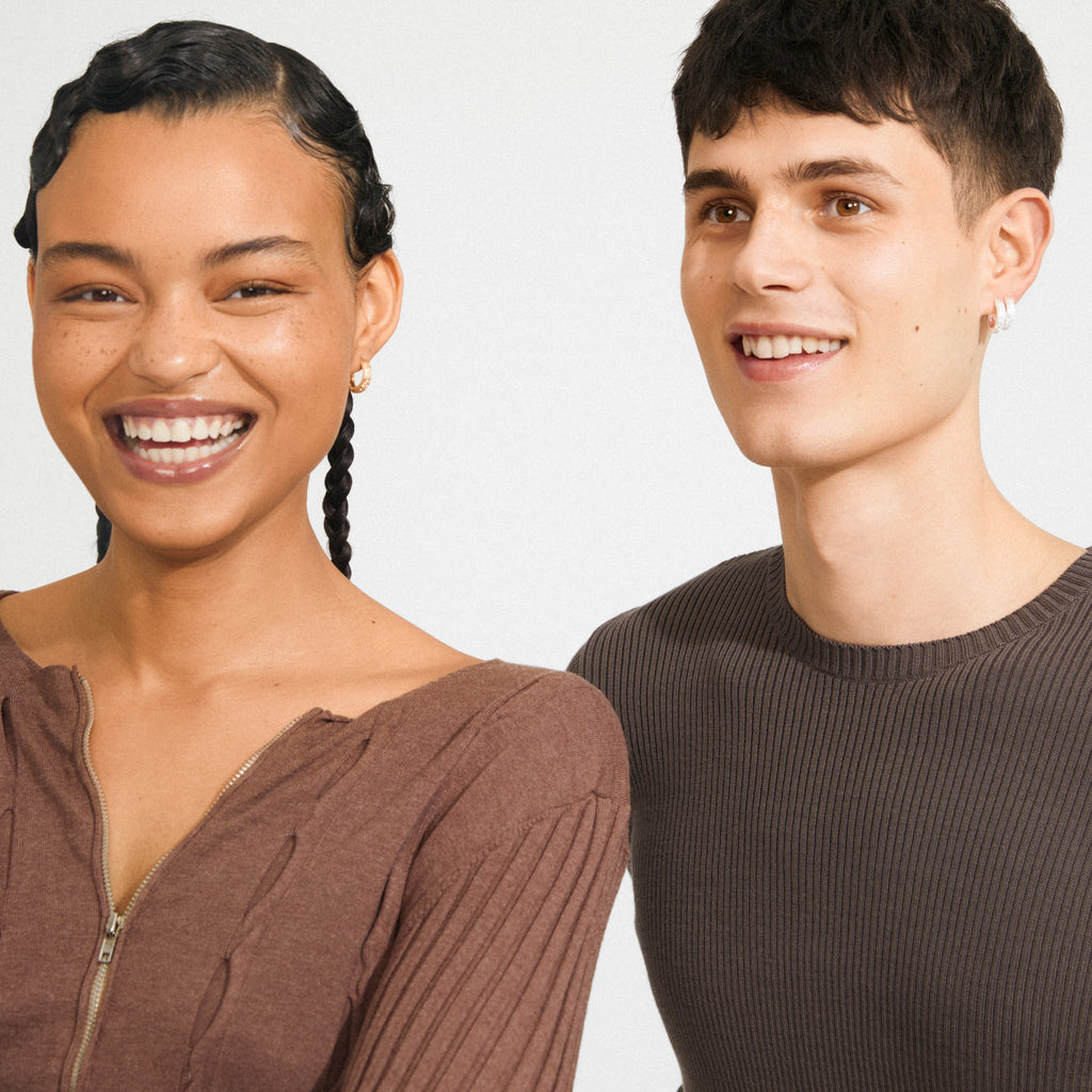 Two young adults showcase ELFRIDA gold-plated hoop earrings, featuring nature-inspired carvings. Both smile warmly, illustrating Pilgrim's blend of timeless and trendy style.