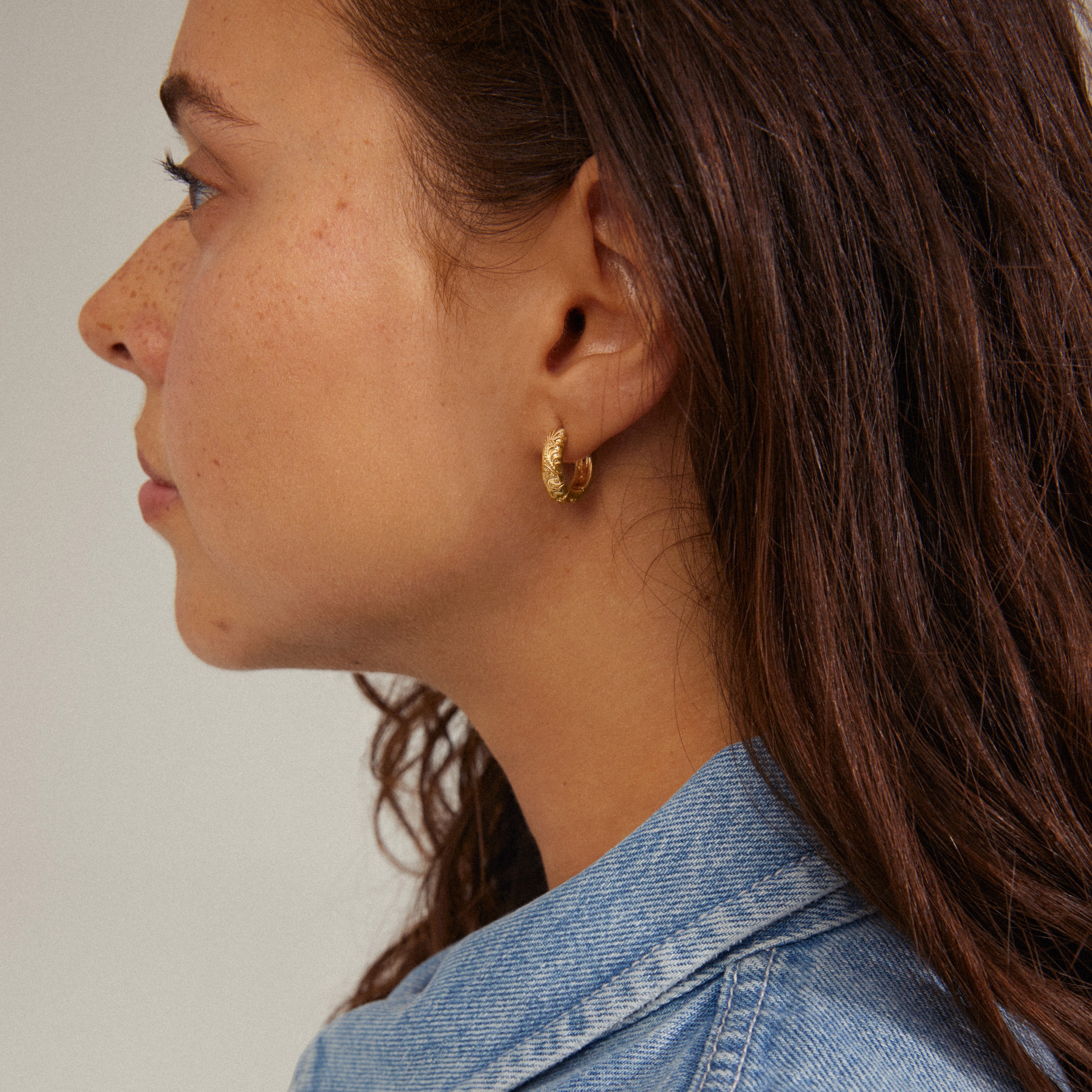 ELFRIDA gold-plated hoop earrings with nature-inspired carvings are shown worn by a woman, highlighting their intricate floral and leaf patterns.