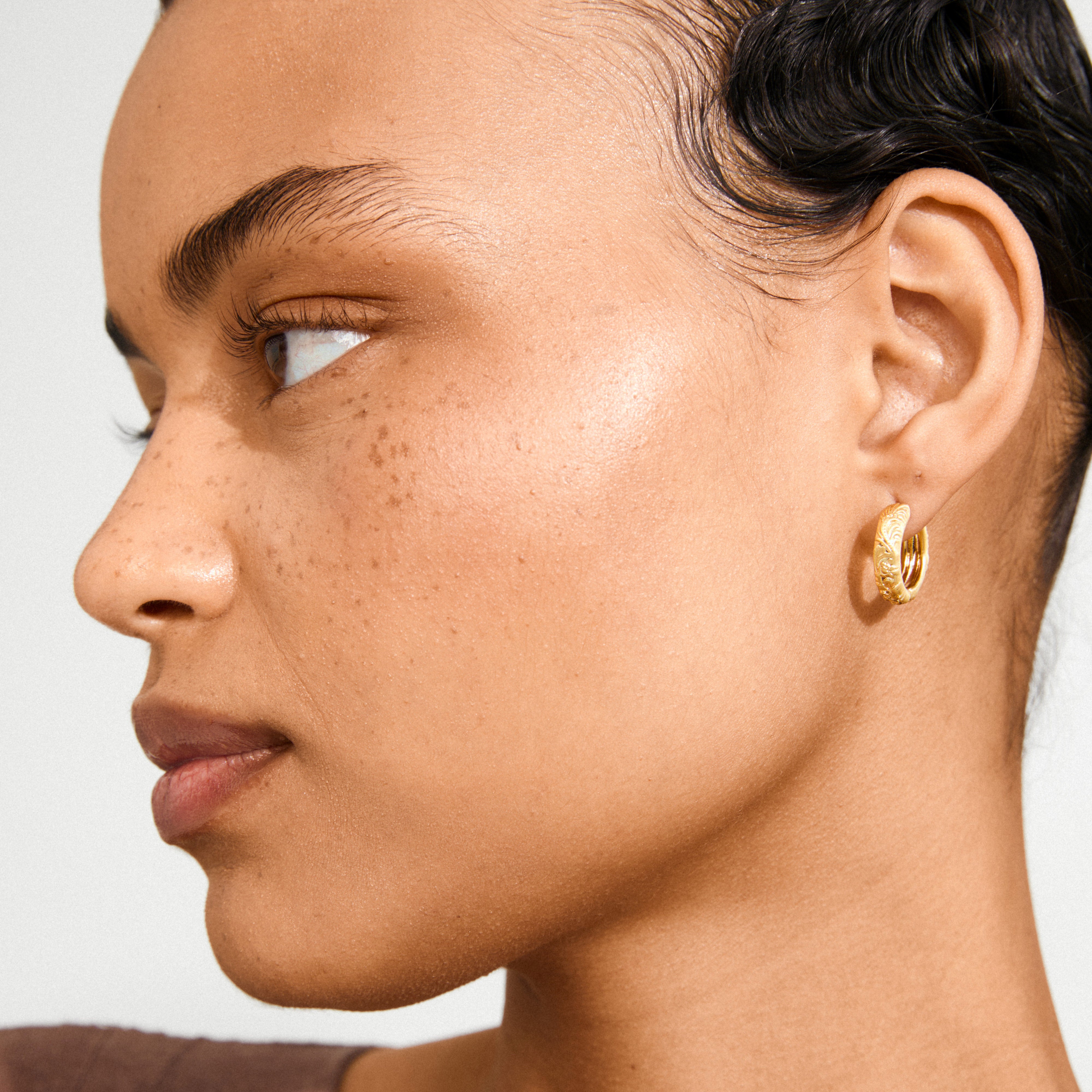 ELFRIDA hoop earrings gold-plated with intricate nature-inspired carvings, worn by a person with slicked-back hair, highlighting the elegant, textured design.