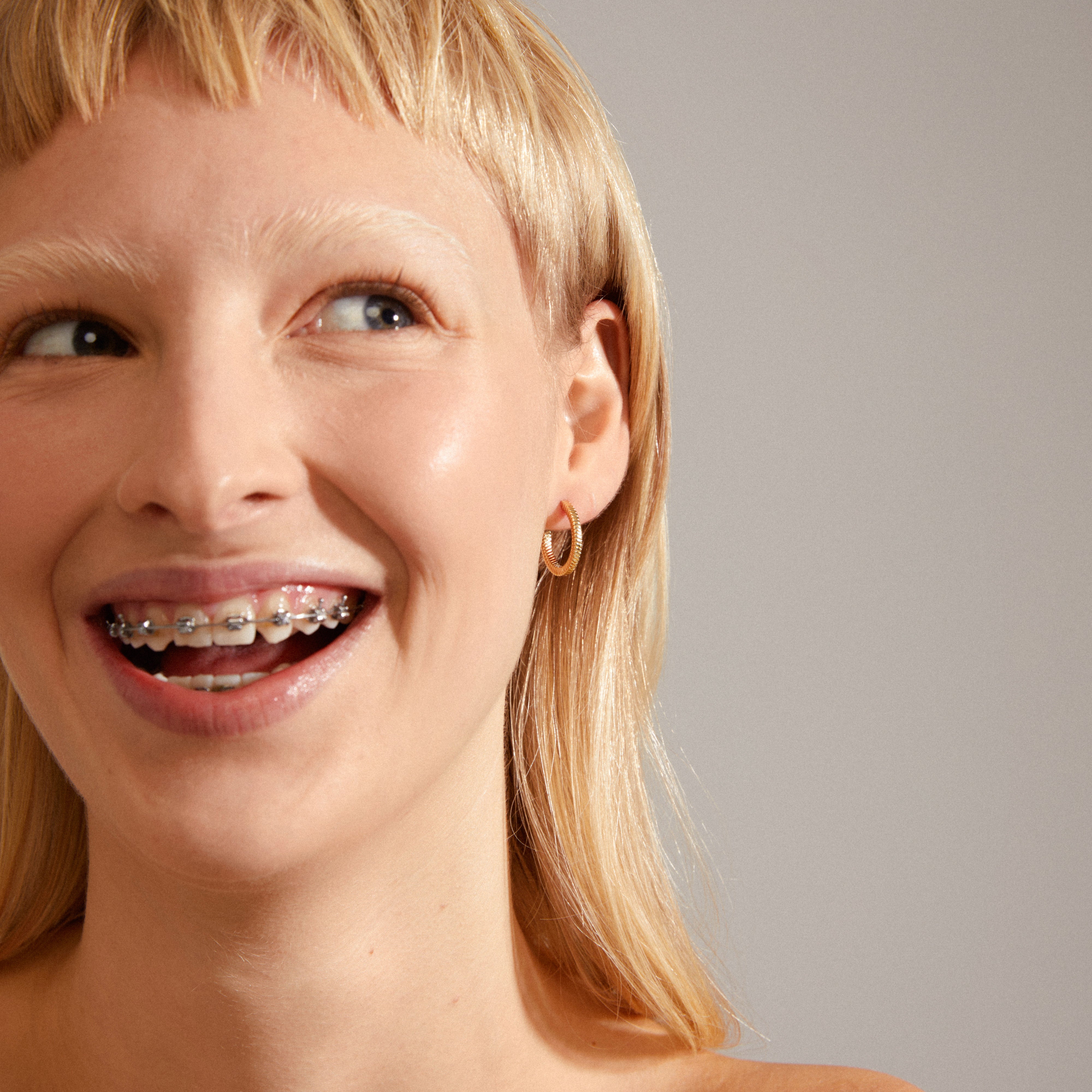 Dominique gold-plated hoop earrings with a unique square snake design and shiny details, worn by a smiling woman showcasing braces.