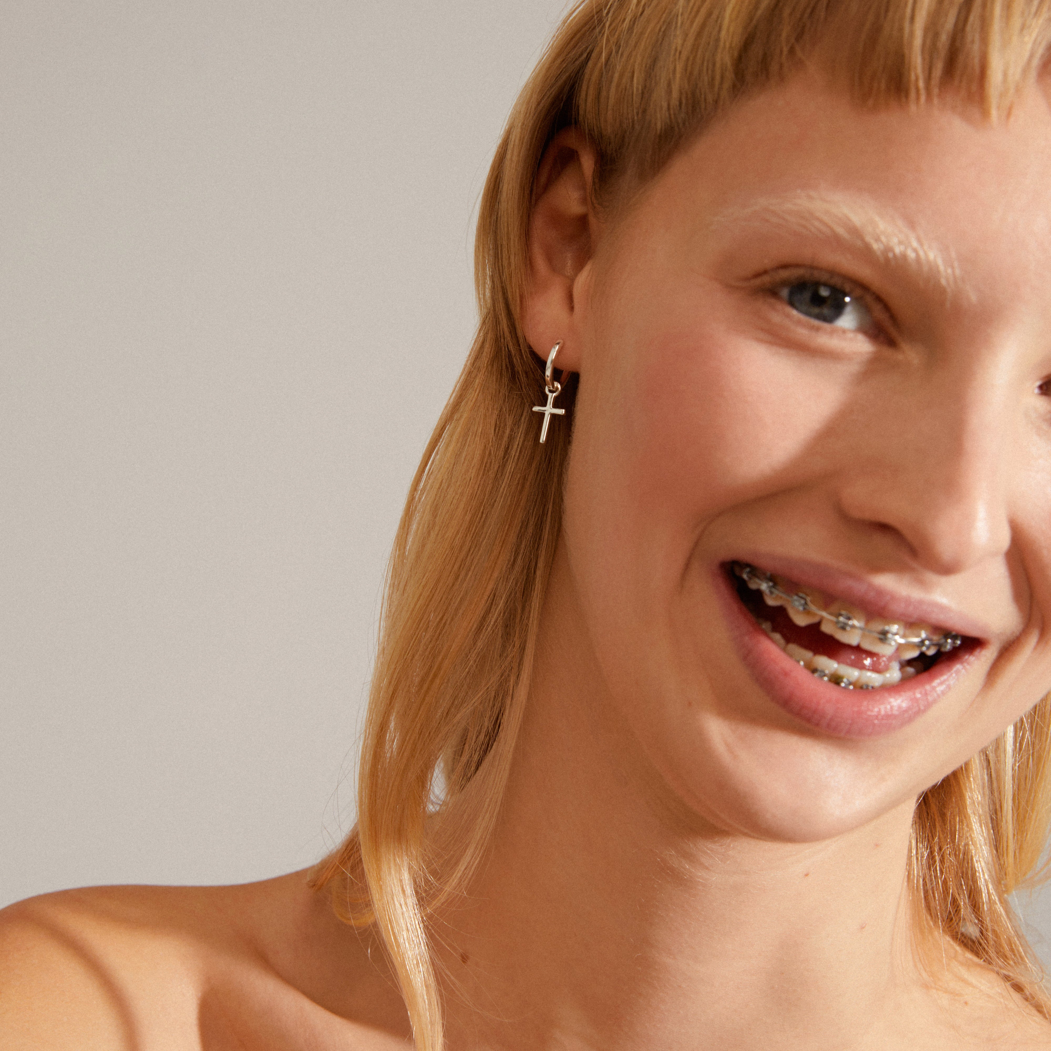 Close-up of a woman smiling, showcasing Pilgrim's DAISY cross hoops, silver-plated huggie earrings with a removable cross pendant for versatile styling.