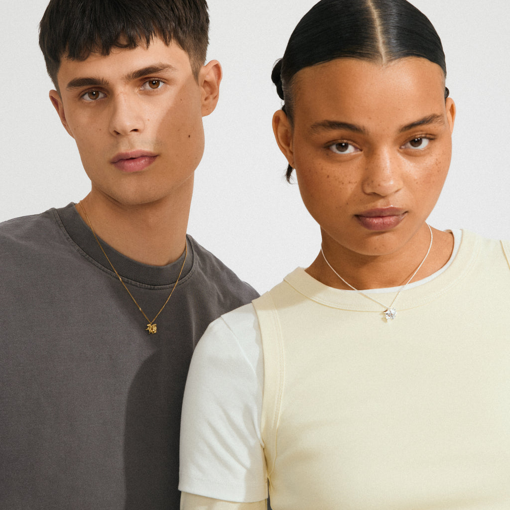 Young man models Pilgrim's gold-plated TIMBER necklace with a delicate flower pendant, showcasing minimalist style and Scandinavian design against a light background.