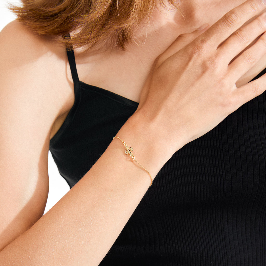 Woman wearing DAGMAR bracelet gold-plated, featuring a delicate gold chain with a flower-like cross charm, resting on her wrist against her collarbone.