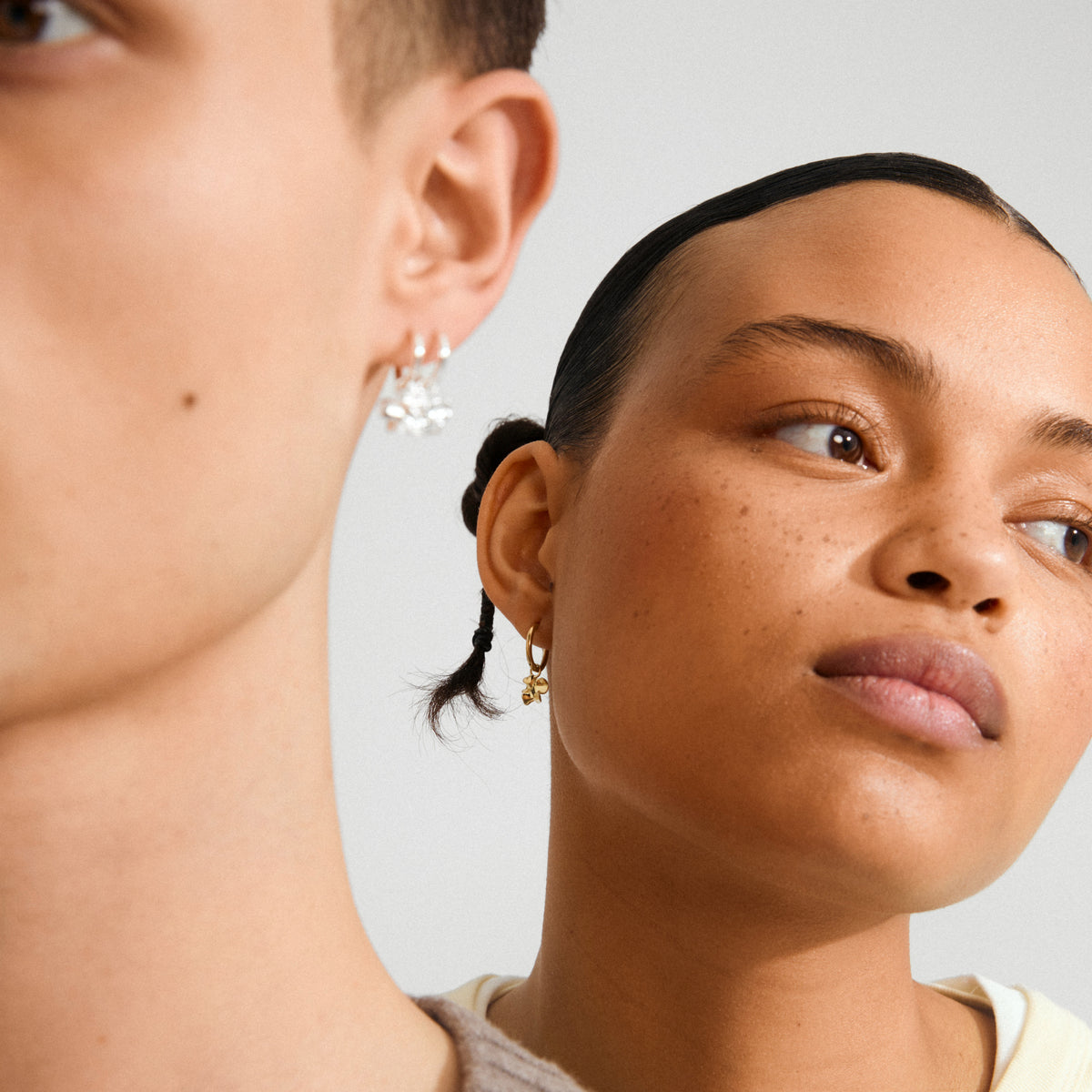 Profile view of two people showcasing Pilgrim's TIMBER earrings: silver with crystal charms and gold-plated with flower charm, highlighting minimalist design and recycled material.
