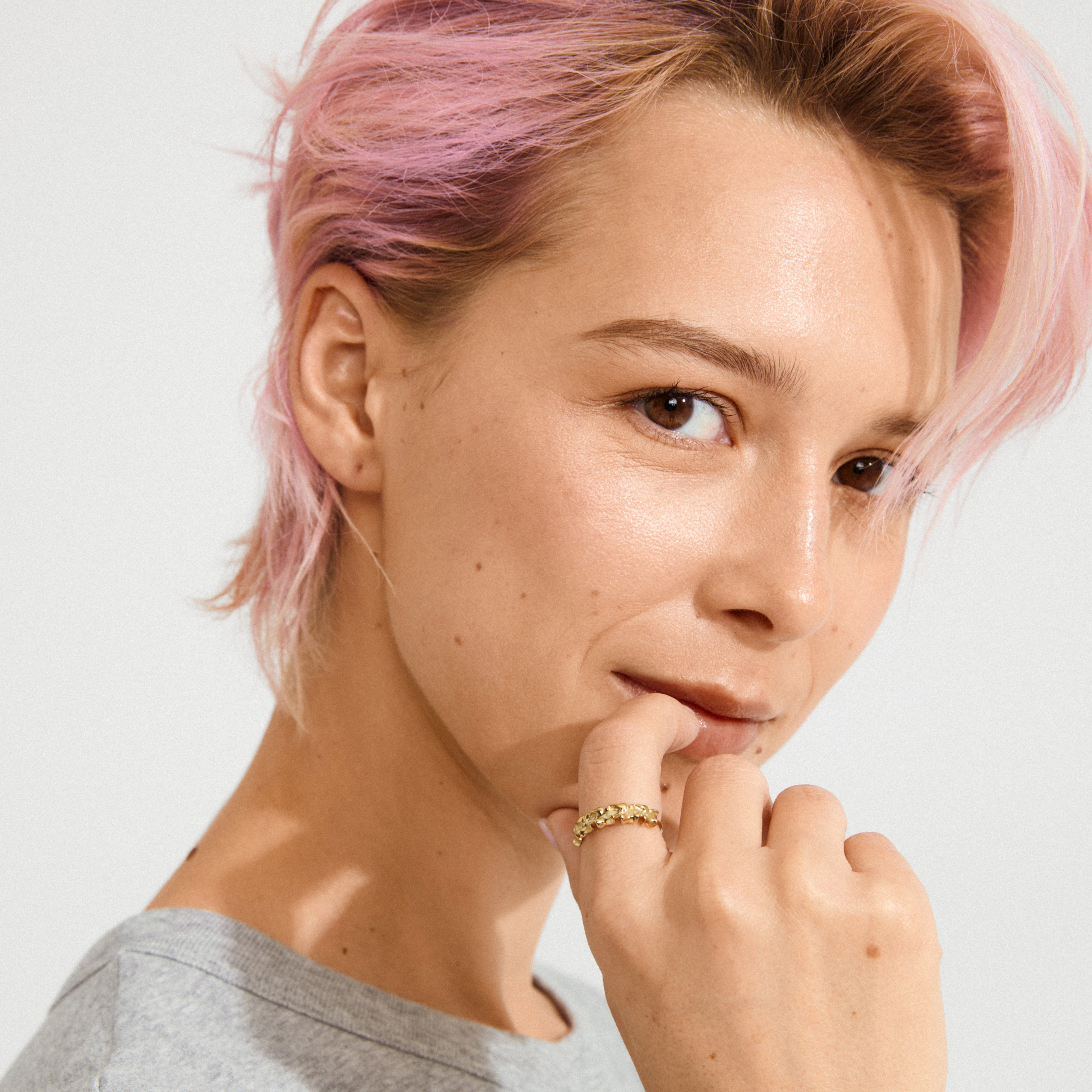 Young adult with pastel-pink hair showcasing the gold-plated TIMBER ring by Pilgrim, featuring abstract floral details, evoking nature's beauty and designed for stacking.