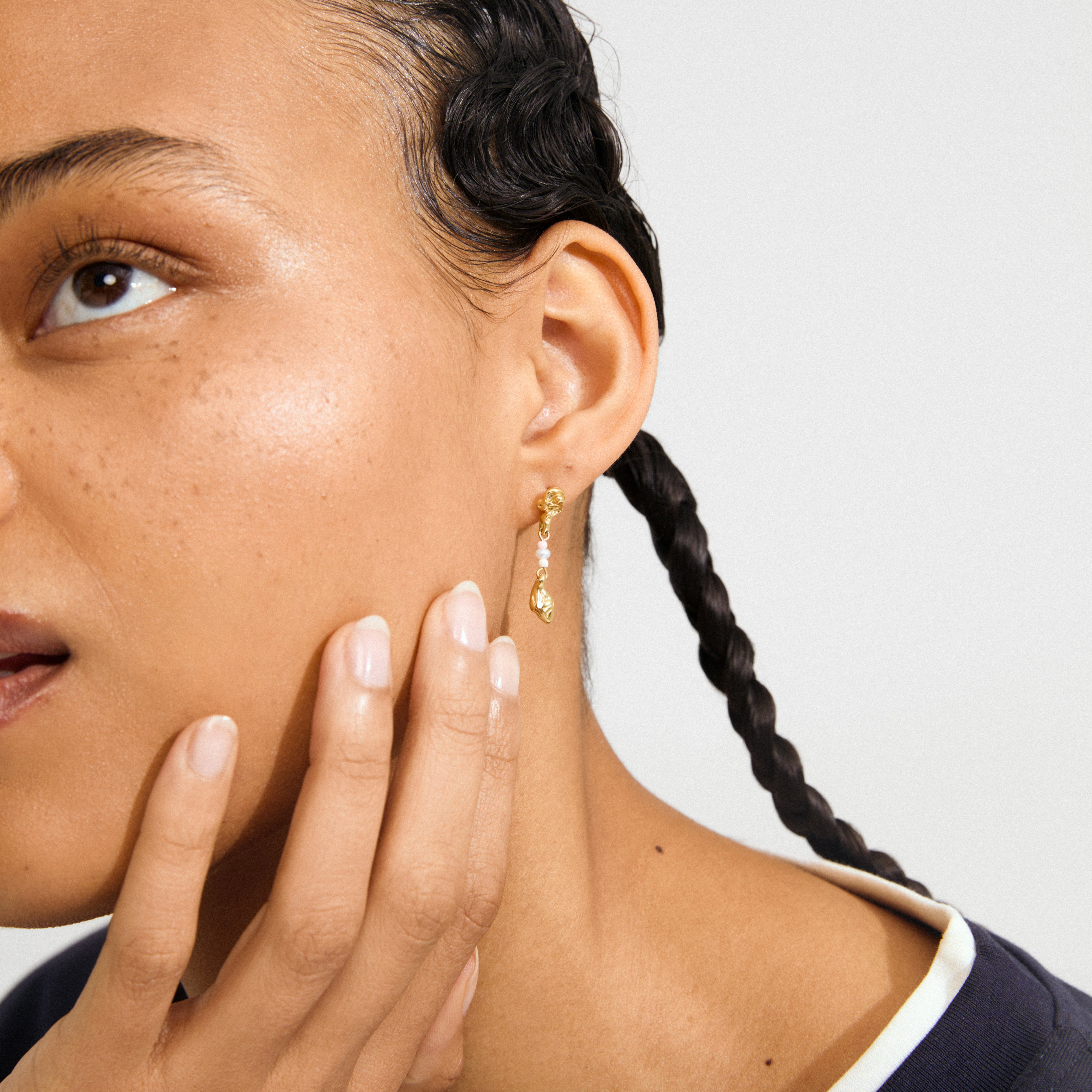 Gold-plated SEQUOIA earring with a white bead detail, worn on a freckled ear, accompanied by manicured hand and braided dark hair.