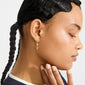 Young person with glowing skin, wearing Pilgrim's gold-plated SEQUOIA earrings featuring textured charm, beads, and pearls, styled with dark braided hair, touching chin.