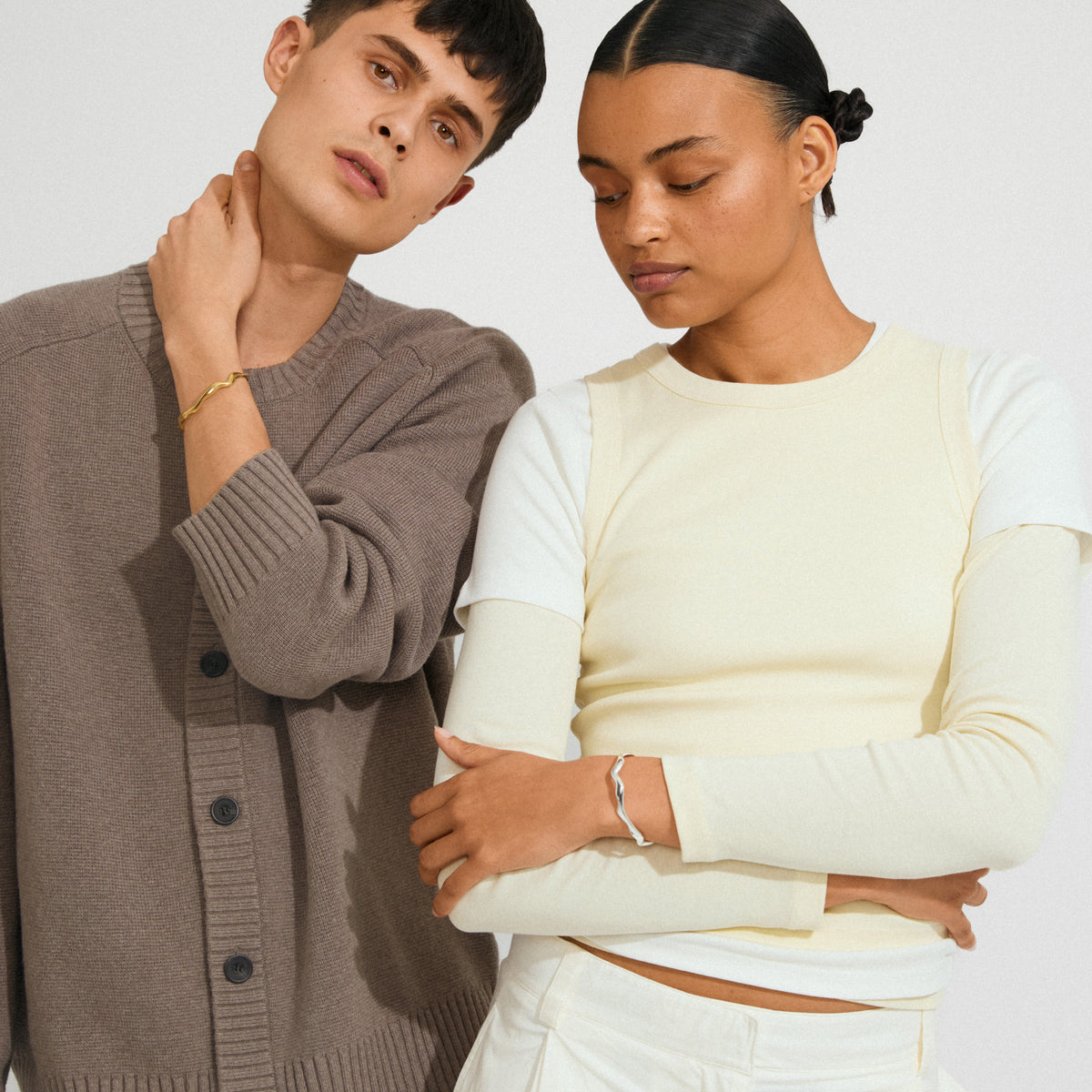 Two models showcase the AIKO bracelet gold-plated by Pilgrim, featuring a wavy, organic design on the young man's wrist in a minimalist fashion setting.