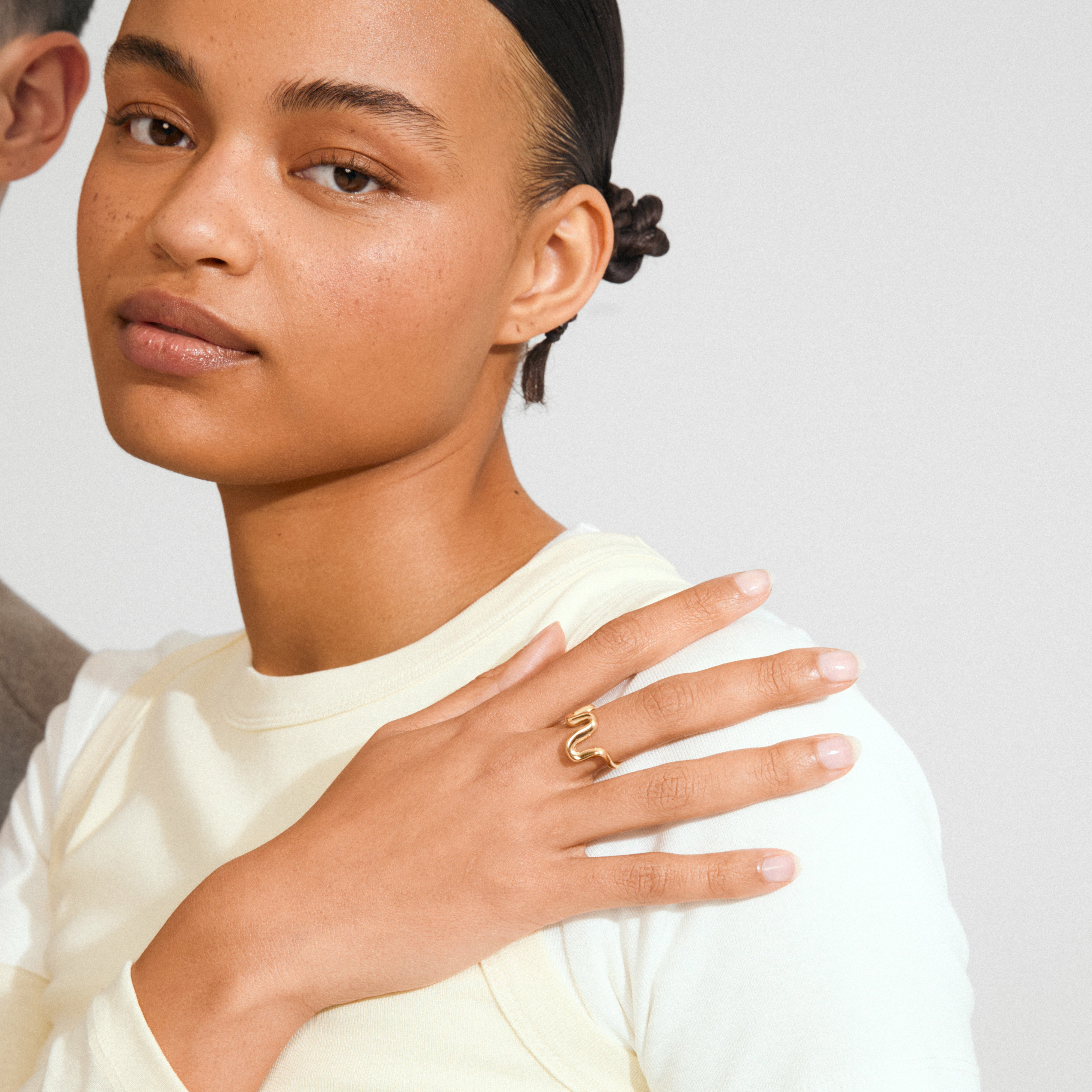 Young woman wearing Pilgrim's AIKO ring, a gold-plated, wavy band on her middle finger, showcasing a sleek, minimalist style with a cream top.