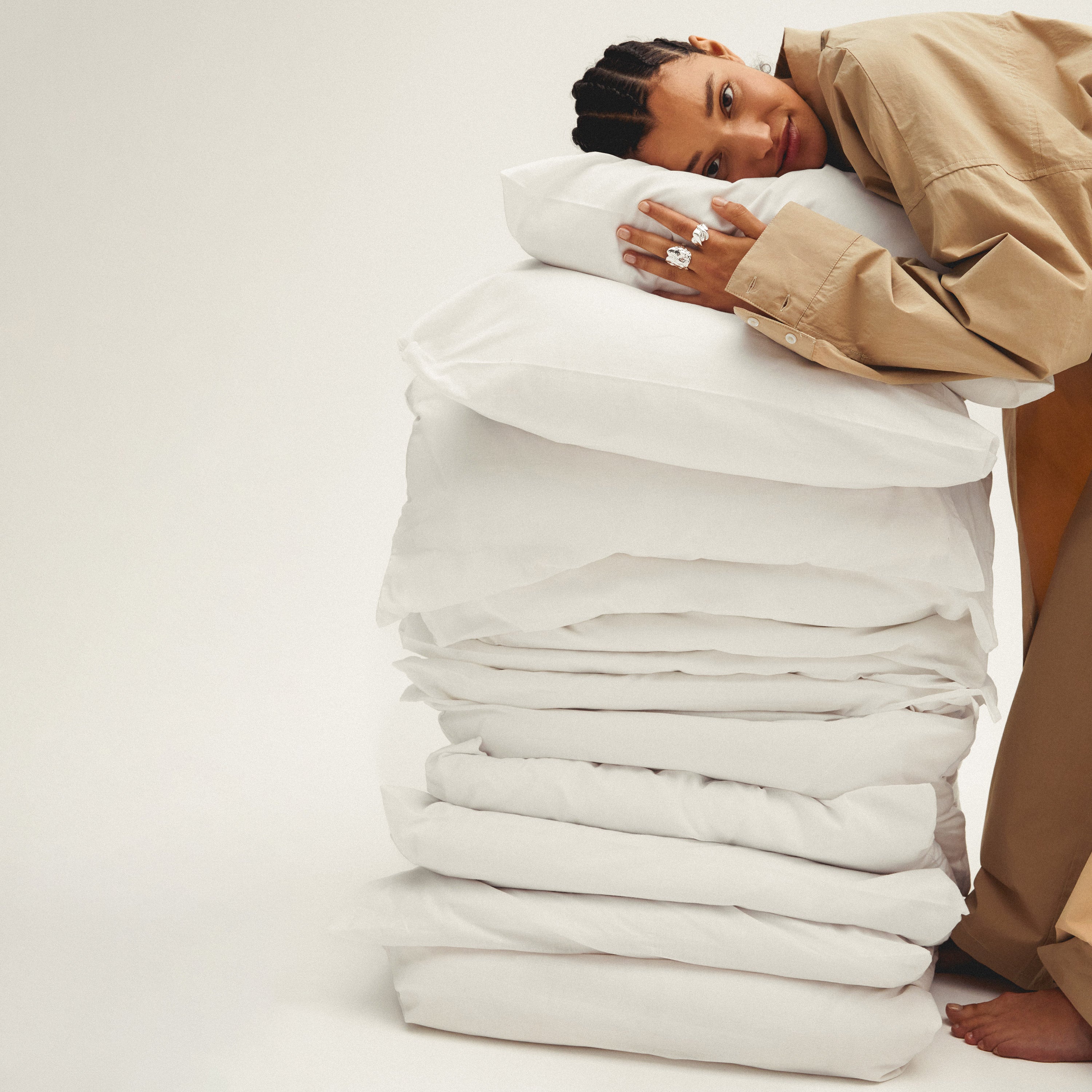 A woman hugging a stack of white pillows, wearing the PILGRIM PJ set XS/S, showcasing its relaxed, oversized fit and cozy style.
