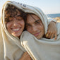 A man and woman wrapped in Pilgrim's large beige beach towel with logo, showcasing its size and soft texture for luxurious comfort.