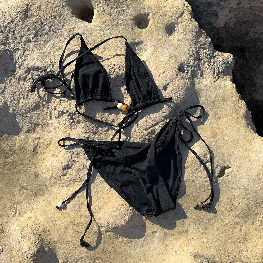 Black bikini with triangle top featuring earthy-tone beads and a tie-neck halter, high-cut bottoms with side ties, in XS/S. Comes in a sleek Pilgrim drawstring pouch.
